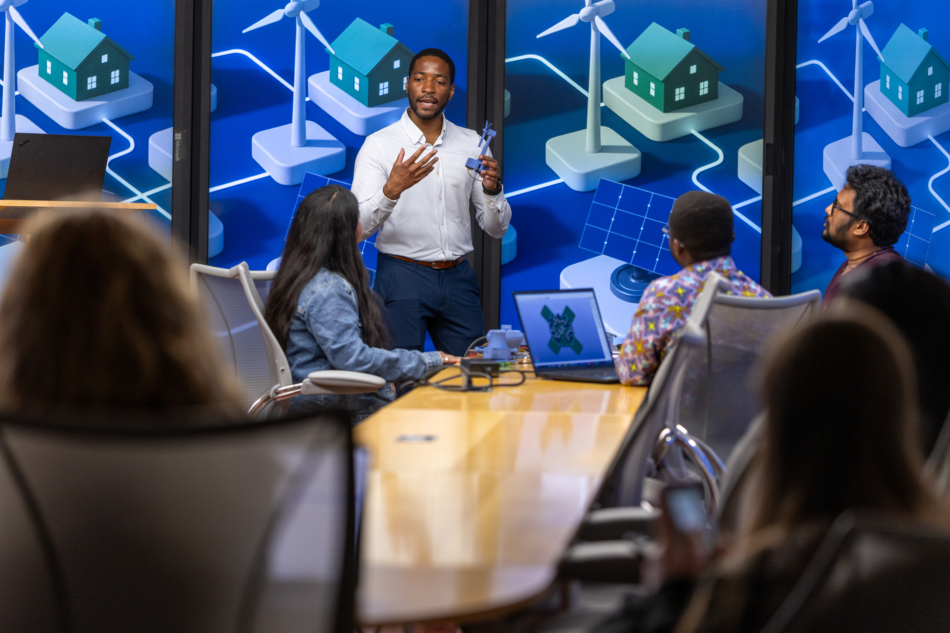 Philip Odonkor standing at the front of a conference room, gesturing with his hands while holding a small model wind turbine, as he speaks to a group of people seated around a long table.