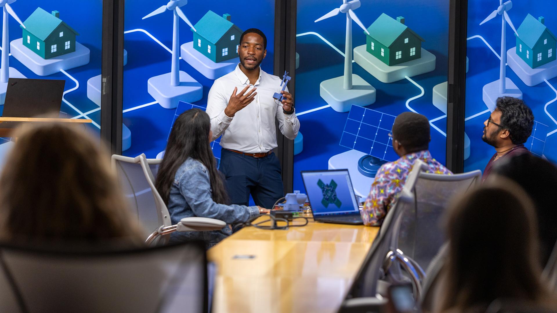 Philip Odonkor standing at the front of a conference room, gesturing with his hands while holding a small model wind turbine, as he speaks to a group of people seated around a long table.