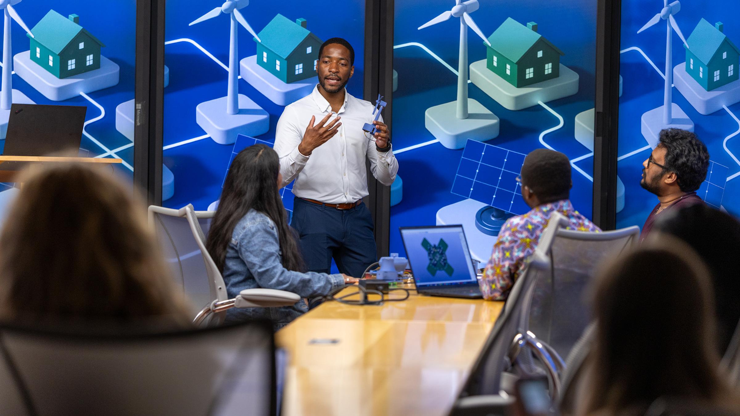 Philip Odonkor standing at the front of a conference room, gesturing with his hands while holding a small model wind turbine, as he speaks to a group of people seated around a long table.