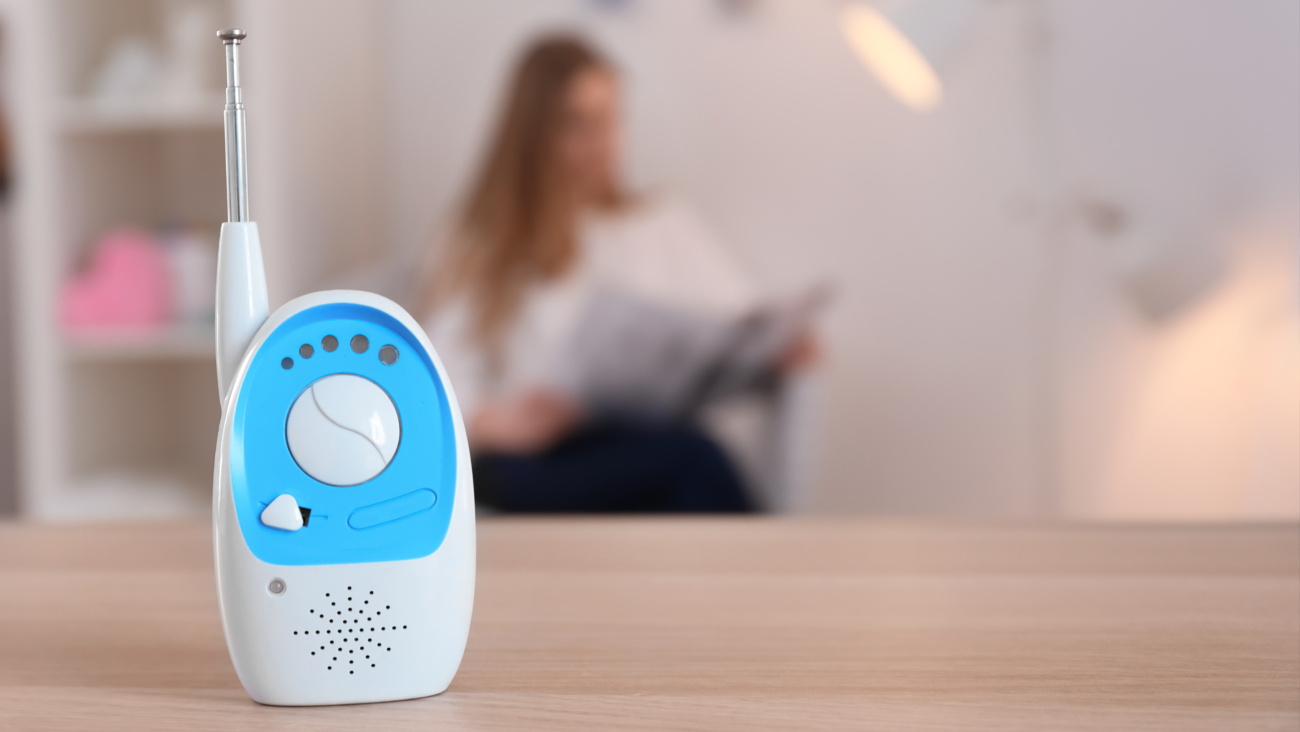A blue and white baby monitor sits on a table in the foreground, against a blurry background showing a person with long blond hair seated reading a magazine.
