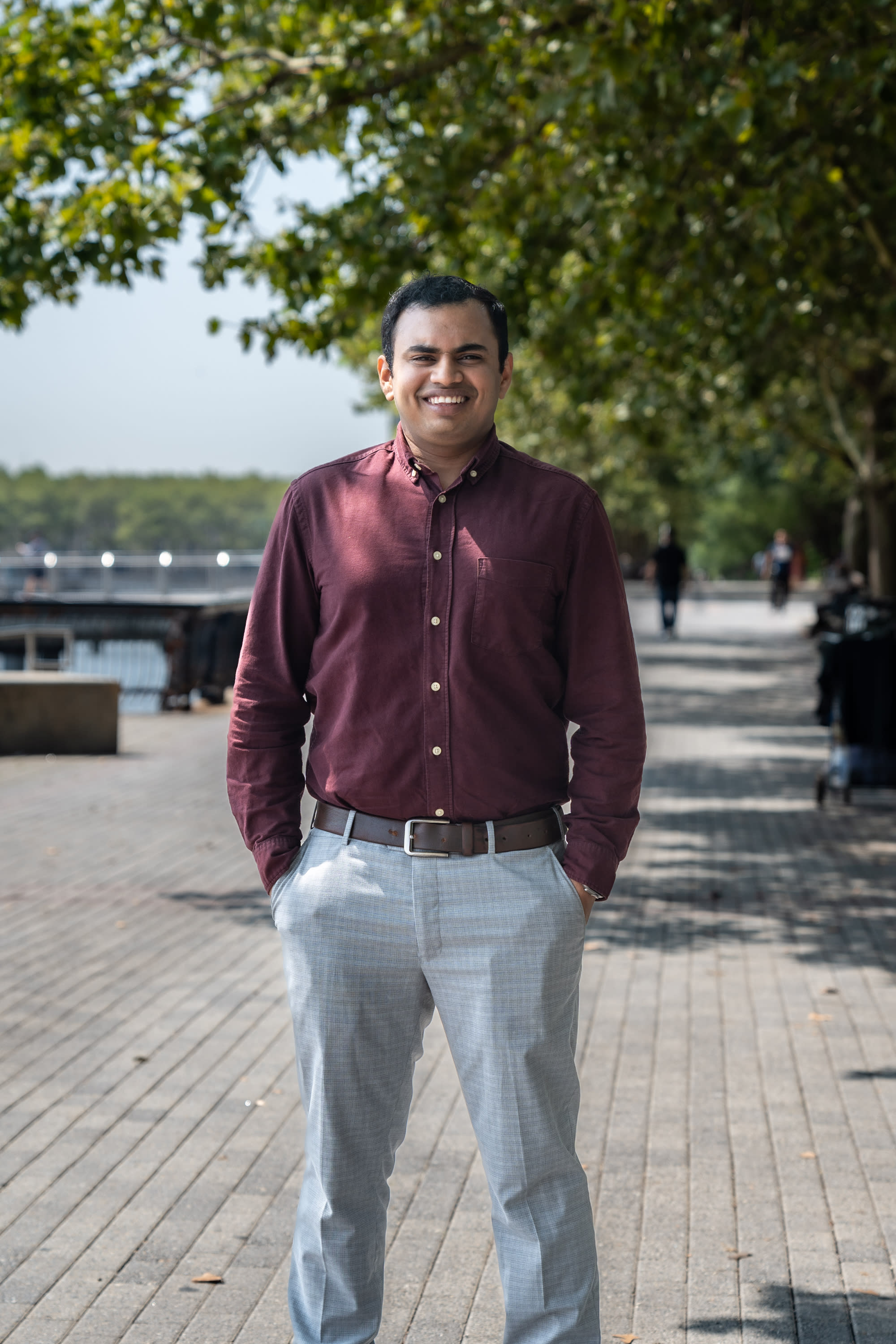 Mani stands with Hoboken's riverwalk in the background on a sunny day. 