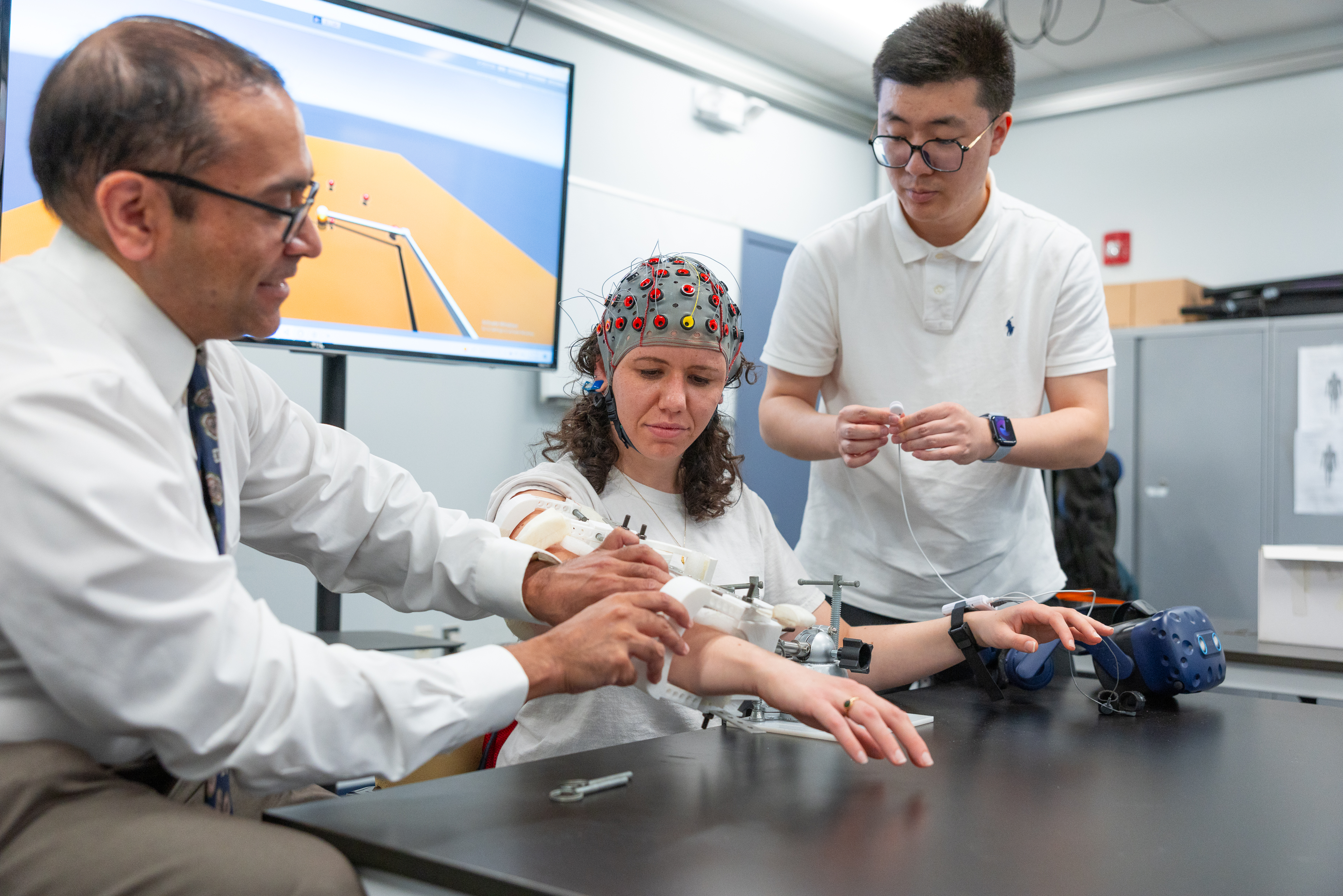 Three people work with medical equipment in a clinical setting; one wears a colorful cap and is seated with their arm extended.