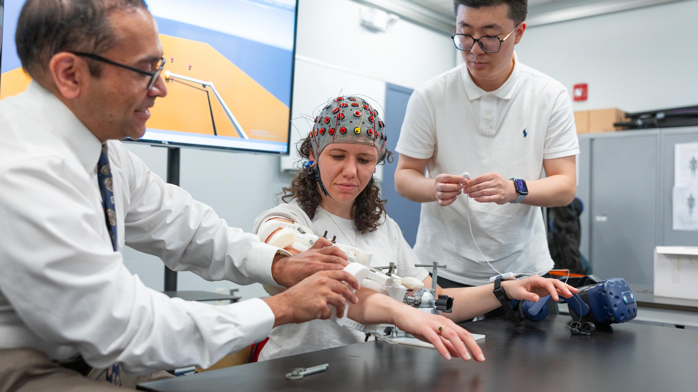 Three people work with medical equipment in a clinical setting; one wears a colorful cap and is seated with their arm extended.