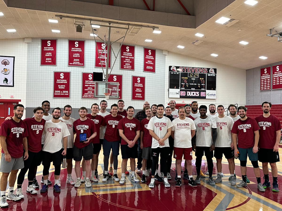Stevens Institute of Technology men’s basketball alumni and current players gather on the court for a group photo during the annual alumni game, standing beneath championship banners and the scoreboard inside Canavan Arena.