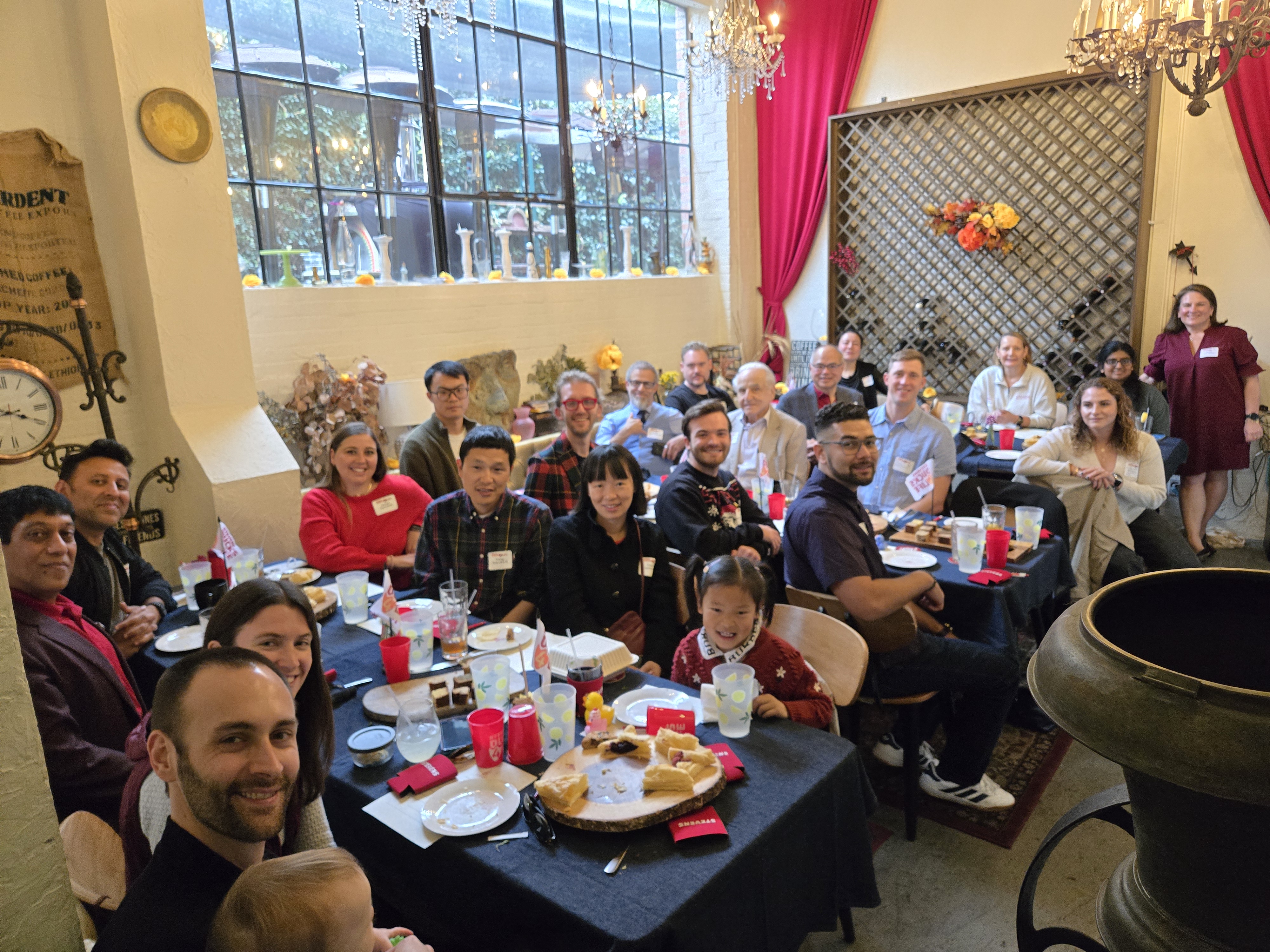 Attendees gather around dining tables at the Southern California Alumni Holiday Gathering 2025, posing for a group photo inside a warmly lit venue with large windows, festive décor and refreshments.