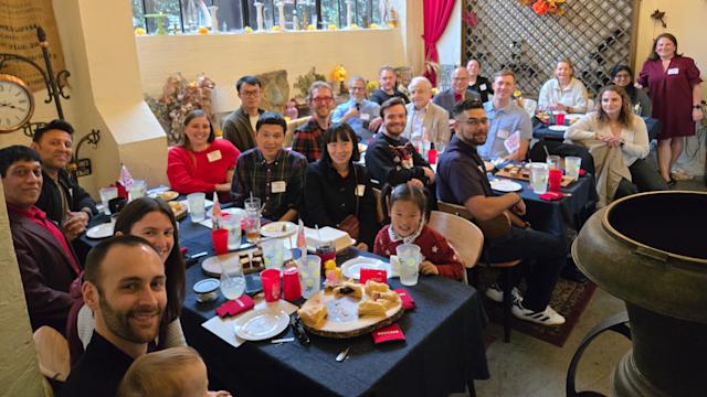 Attendees gather around dining tables at the Southern California Alumni Holiday Gathering 2025, posing for a group photo inside a warmly lit venue with large windows, festive décor and refreshments.