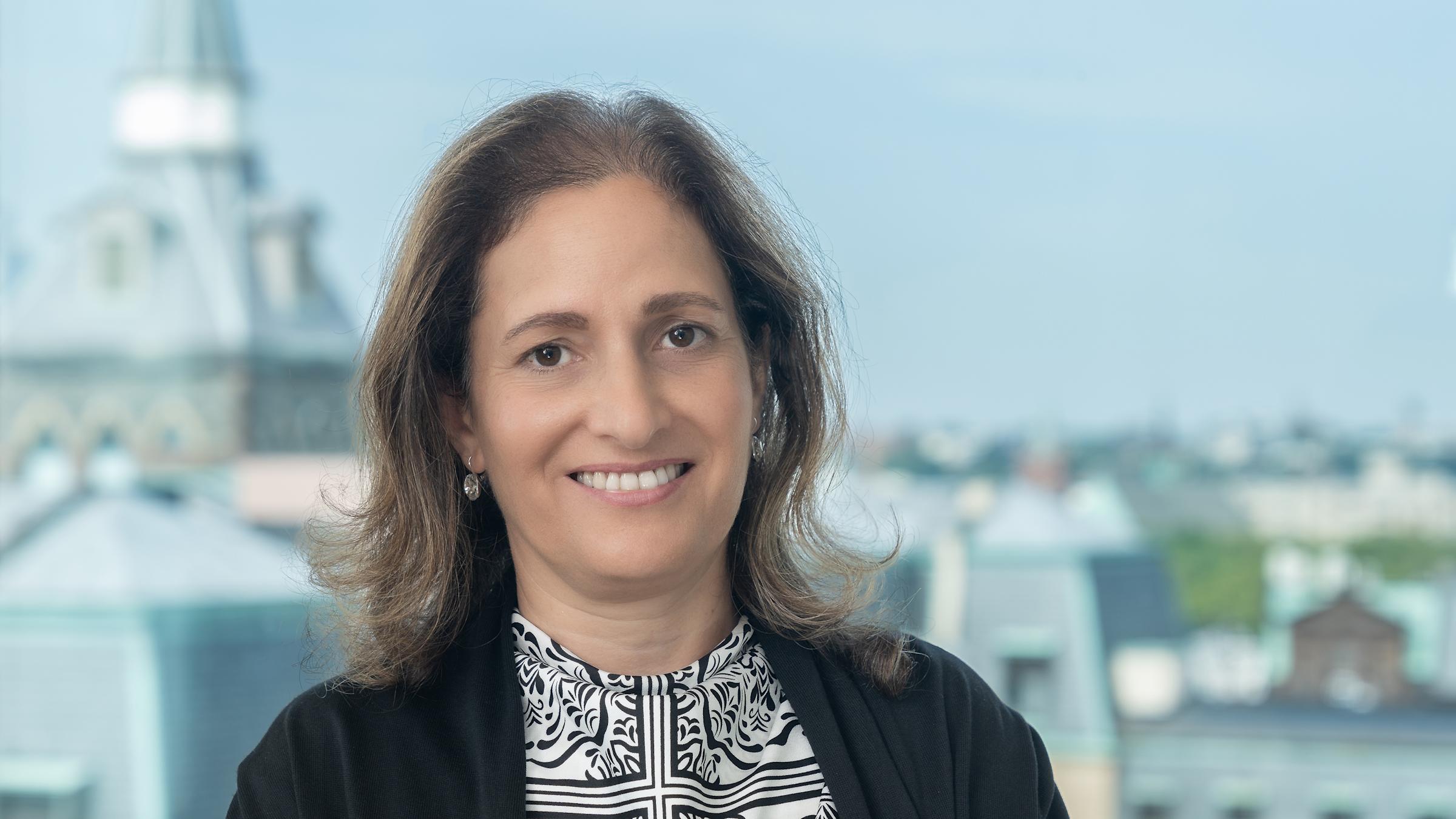 Dr. Joelle Saad-Lessler stands in front of a window with the New York City skyline in the background.