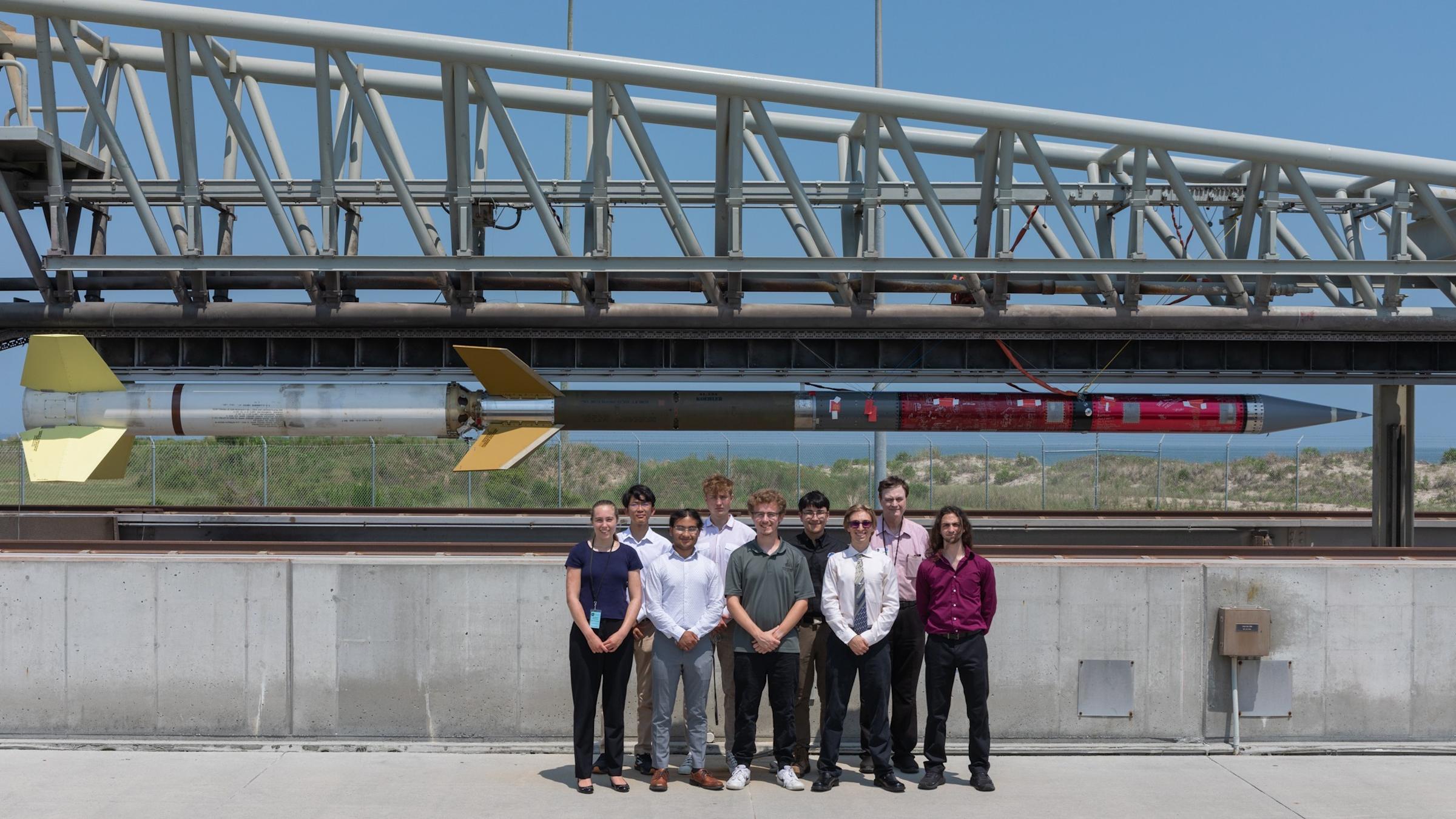 Nine Stevens students stand in front of a concrete partition. Behind them is a bridge. Suspended below the bridge is a rocket spanning the width of the photo.