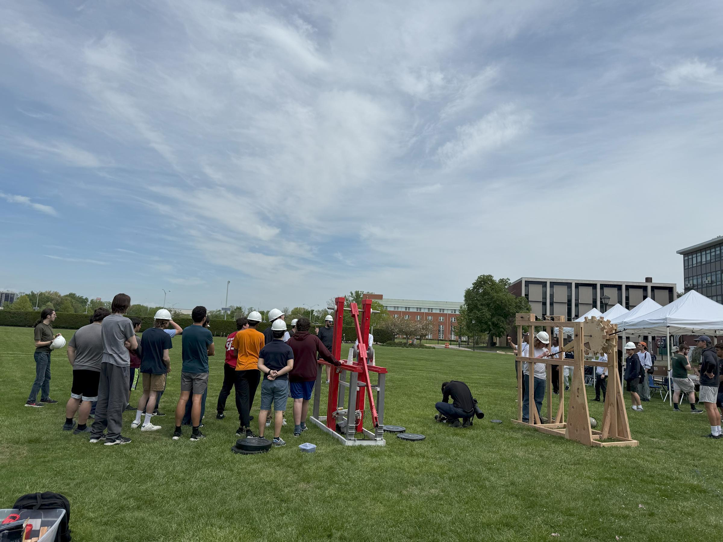A group of people stand with their backs to the camera, wearing hard hats, next to a wooden trebuchet 