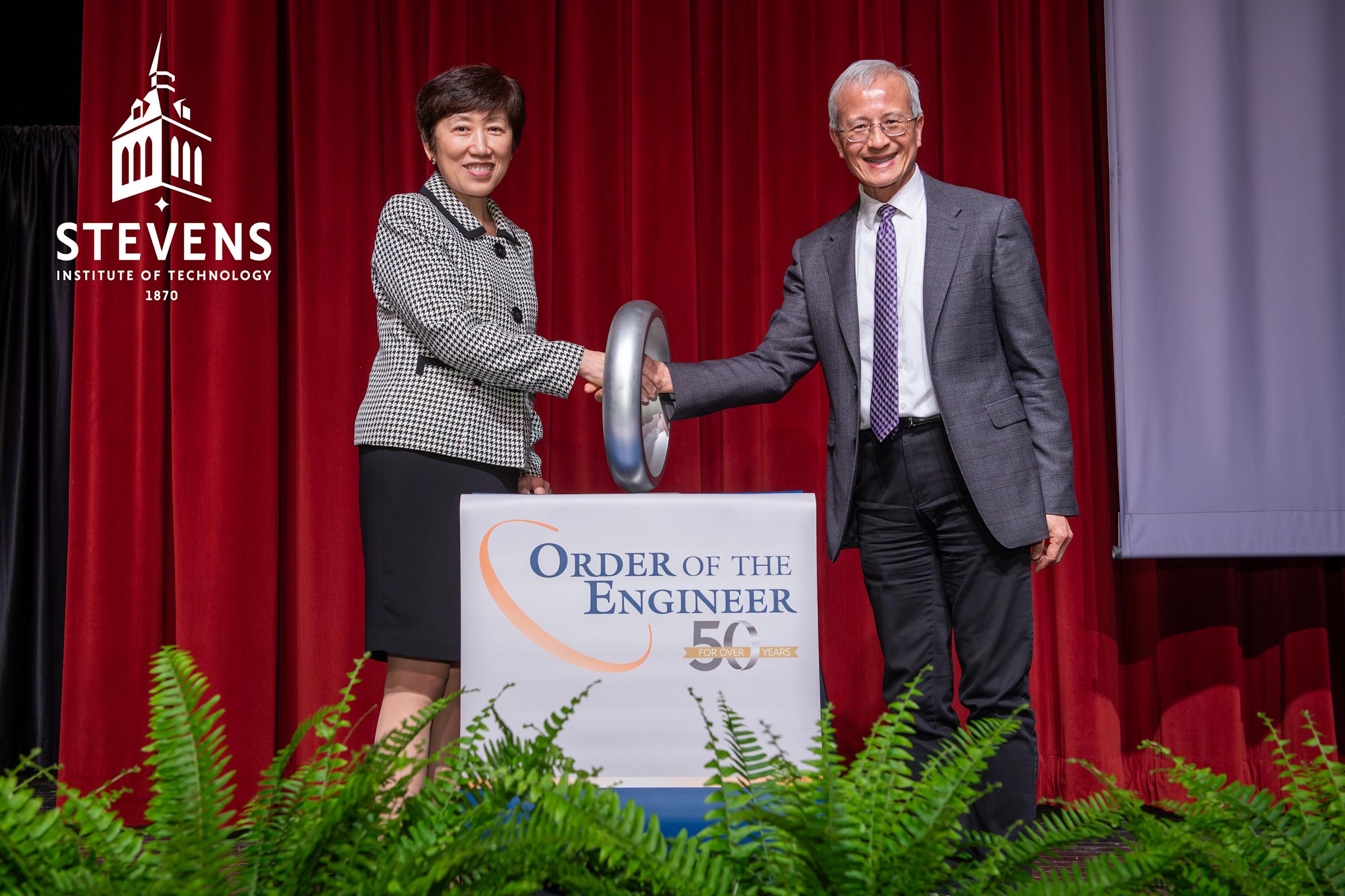 Two people stand behind a podium wearing business clothes. On the podium it reads "Order of the Engineer." The two people are shaking hands through a large ring that is on the podium.