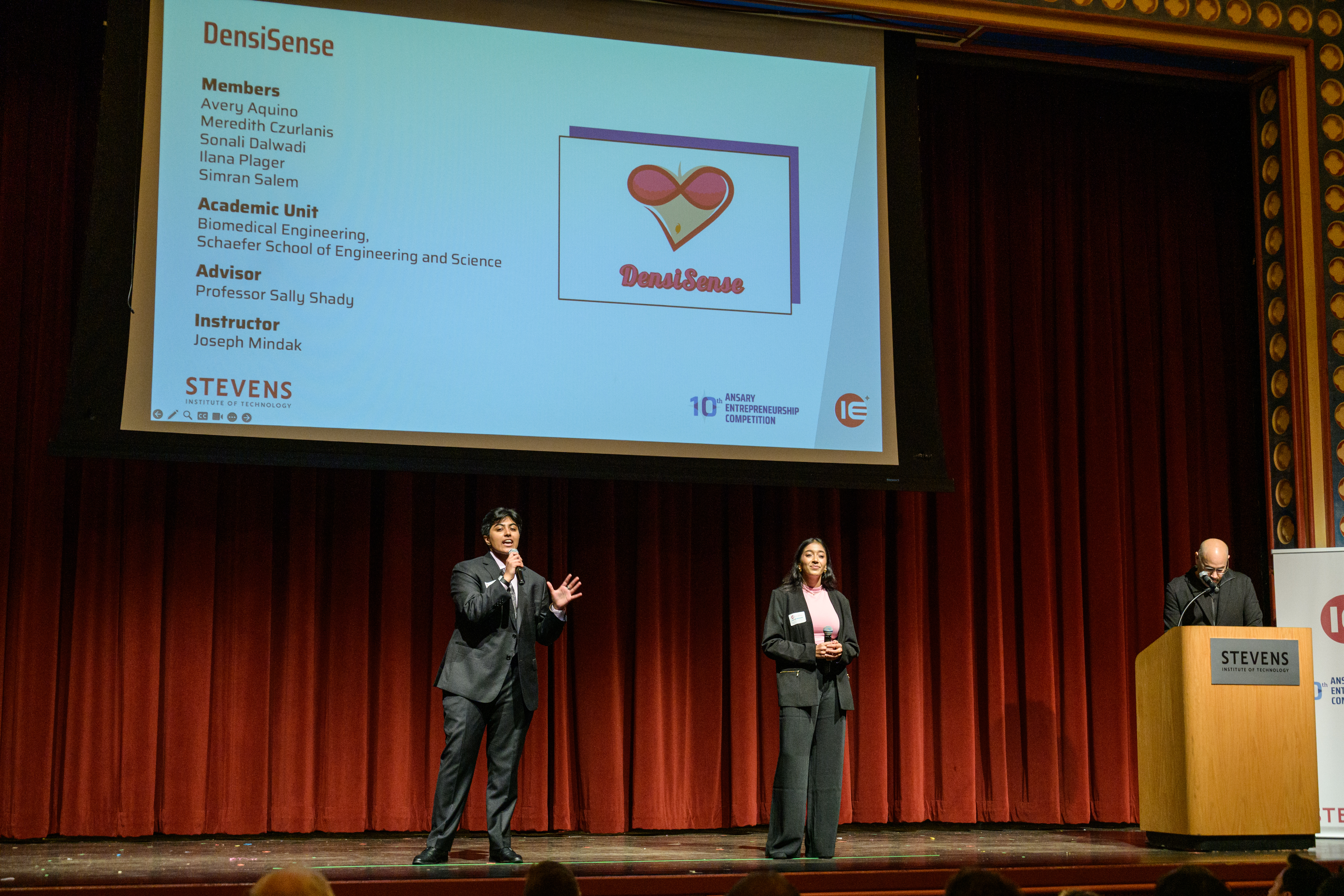 Simran Salem and Sonali Dalwadi talking into wireless microphones in front of a presentation screen detailing their project DensiSense on stage at the 2025 Ansary Competition.