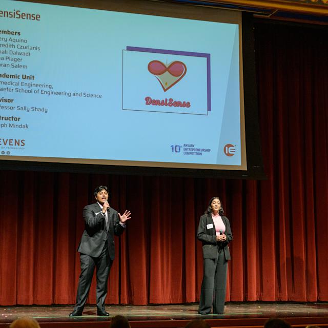 Simran Salem and Sonali Dalwadi talking into wireless microphones in front of a presentation screen detailing their project DensiSense on stage at the 2025 Ansary Competition.