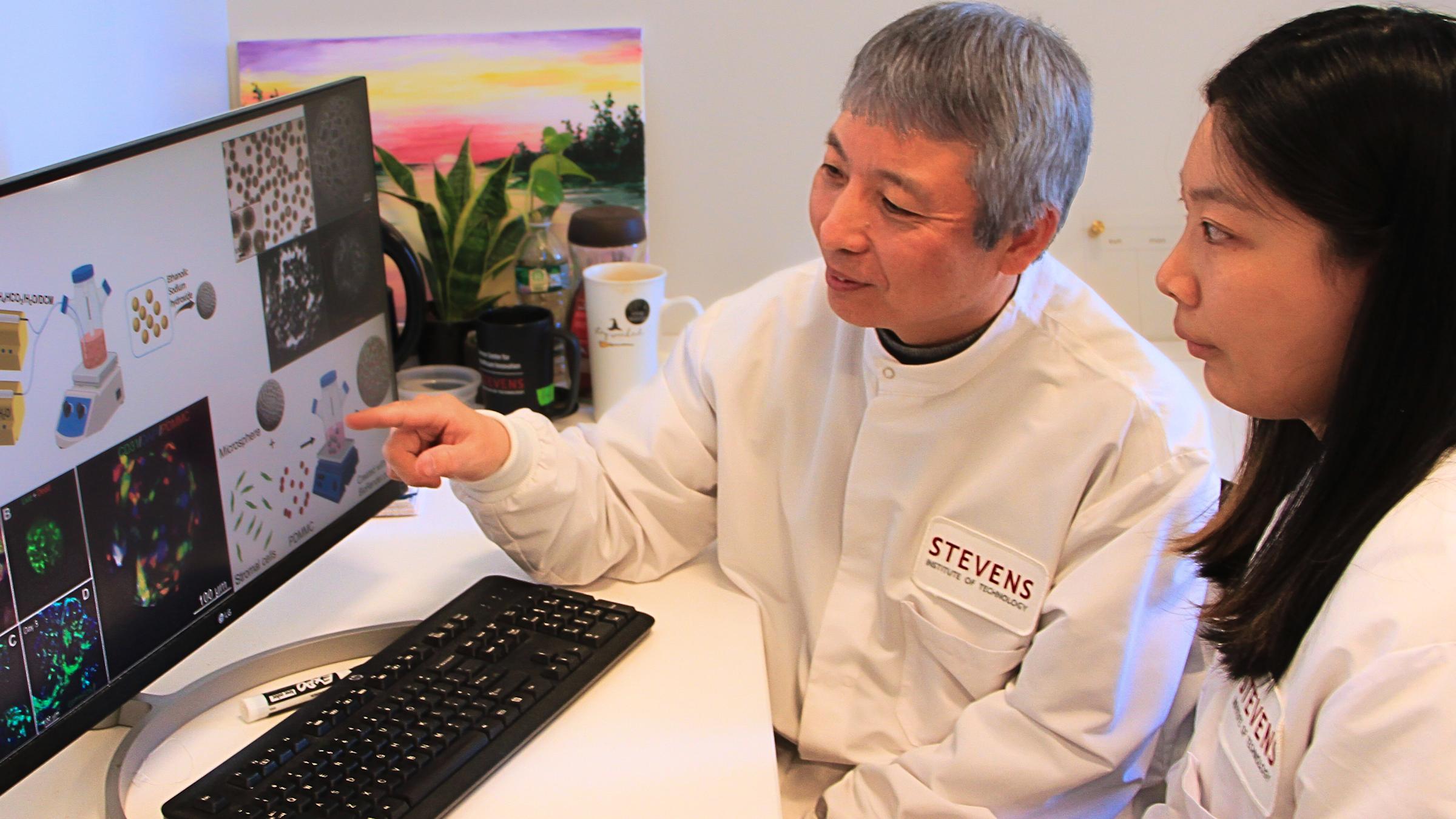 A man with gray hair points at a computer monitor with a woman with black hair to his left.