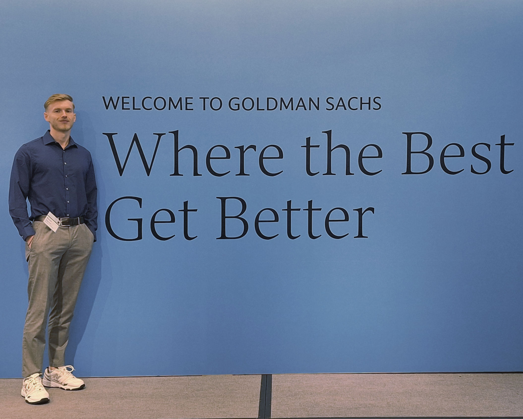 "Young professional in blue shirt and khakis stands casually against Goldman Sachs wall reading 'Welcome to Goldman Sachs - Where the Best Get Better'"