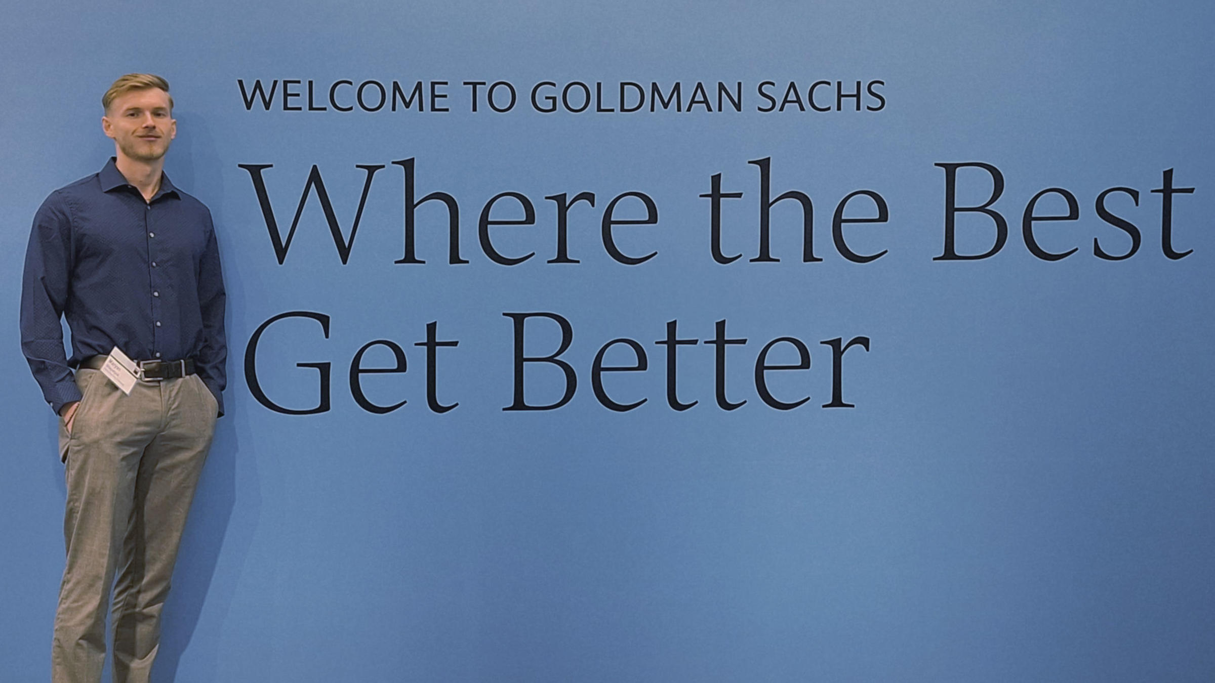 "Young professional in blue shirt and khakis stands casually against Goldman Sachs wall reading 'Welcome to Goldman Sachs - Where the Best Get Better'"