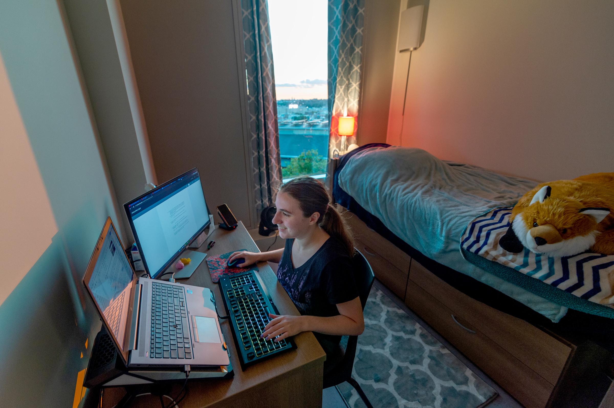Student cheerfully types on her laptop while stuffed fox looks on.