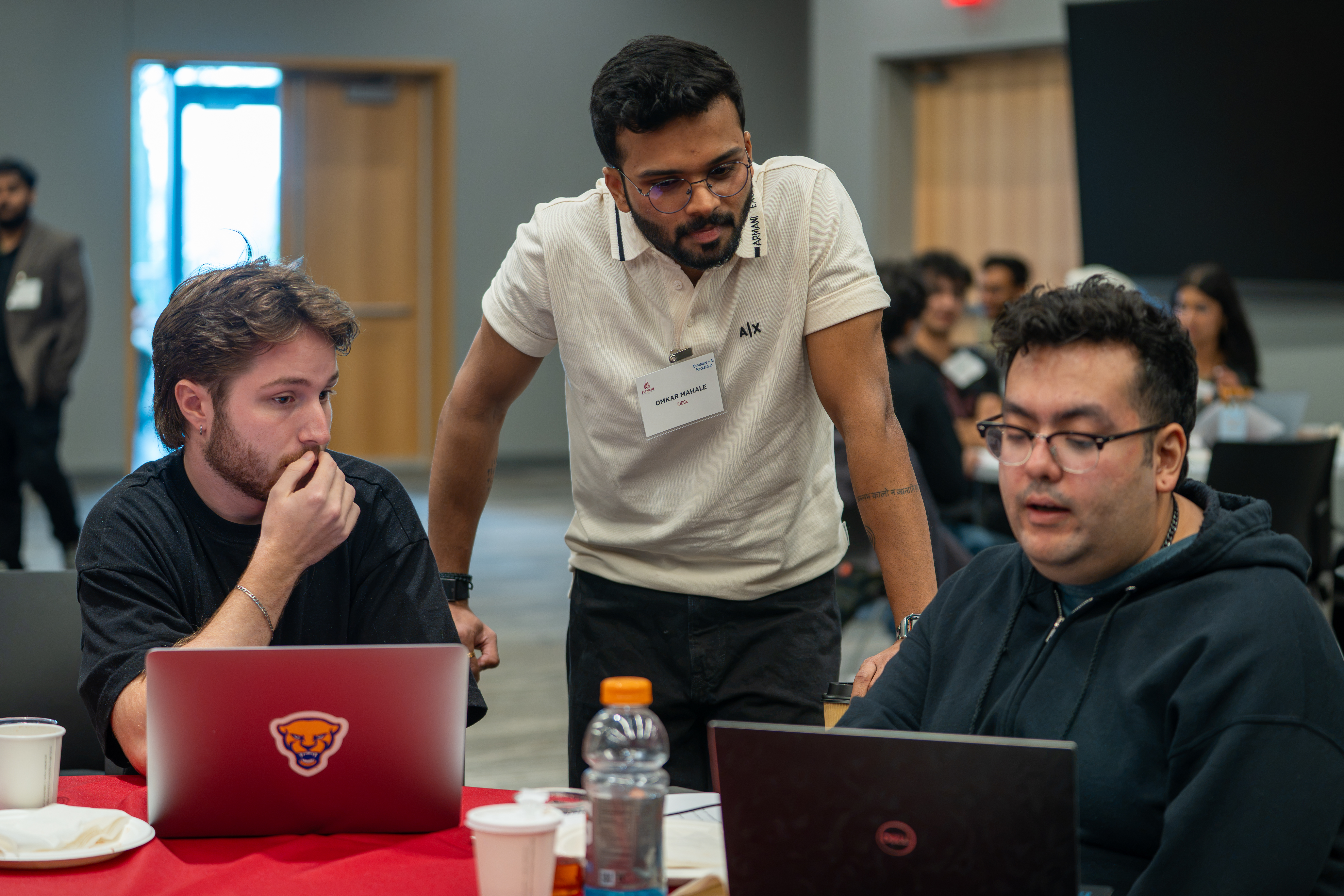 Hackathon judge Omkar Mahale, wearing a cream polo and name badge, leans in to review a project with two seated participants at their laptops on a red-draped table during the Stevens Business + AI Hackathon.