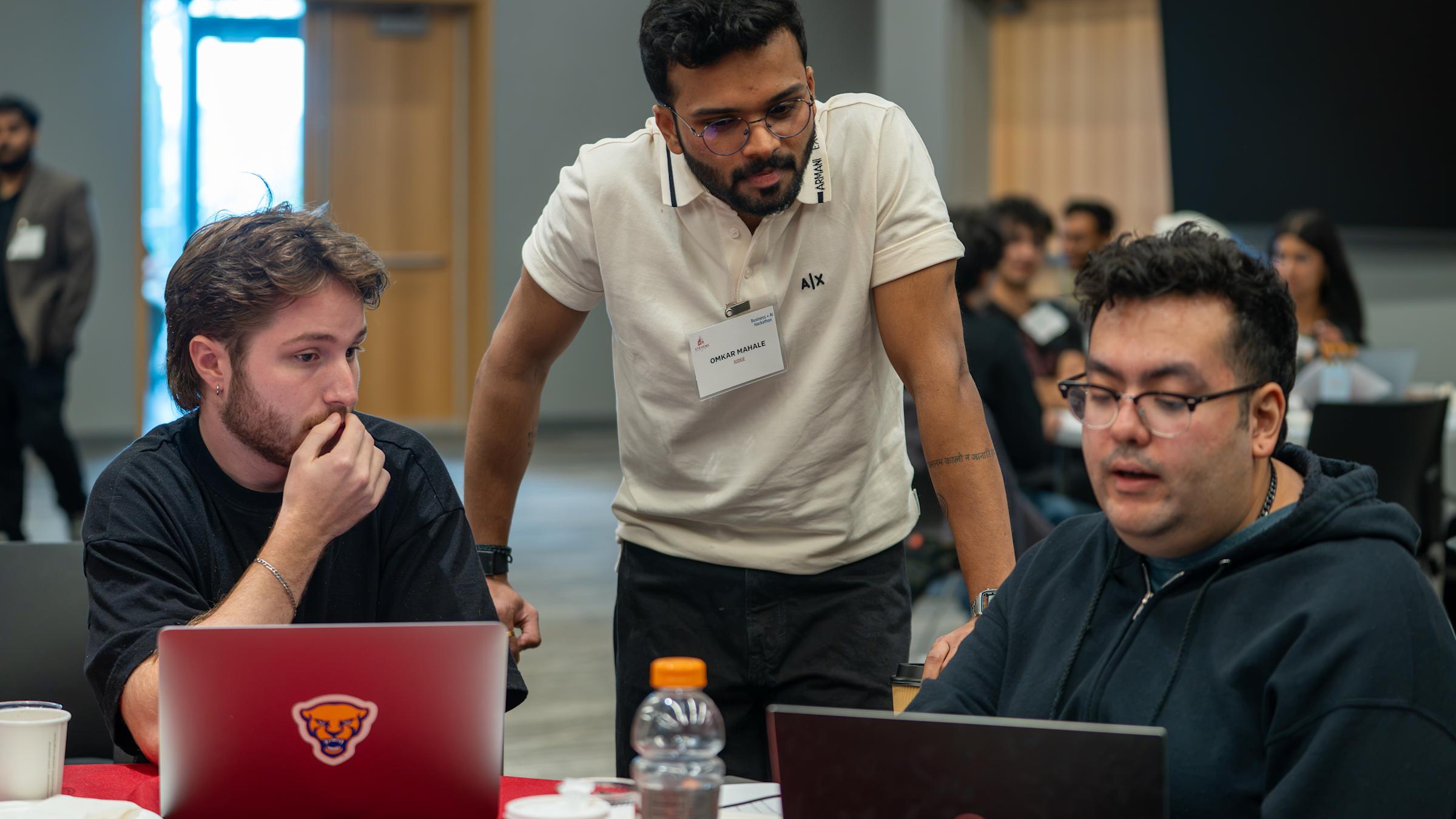 Hackathon judge Omkar Mahale, wearing a cream polo and name badge, leans in to review a project with two seated participants at their laptops on a red-draped table during the Stevens Business + AI Hackathon.