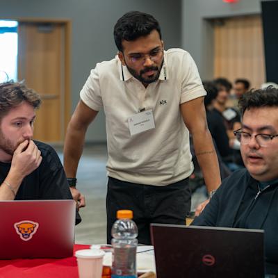 Hackathon judge Omkar Mahale, wearing a cream polo and name badge, leans in to review a project with two seated participants at their laptops on a red-draped table during the Stevens Business + AI Hackathon.