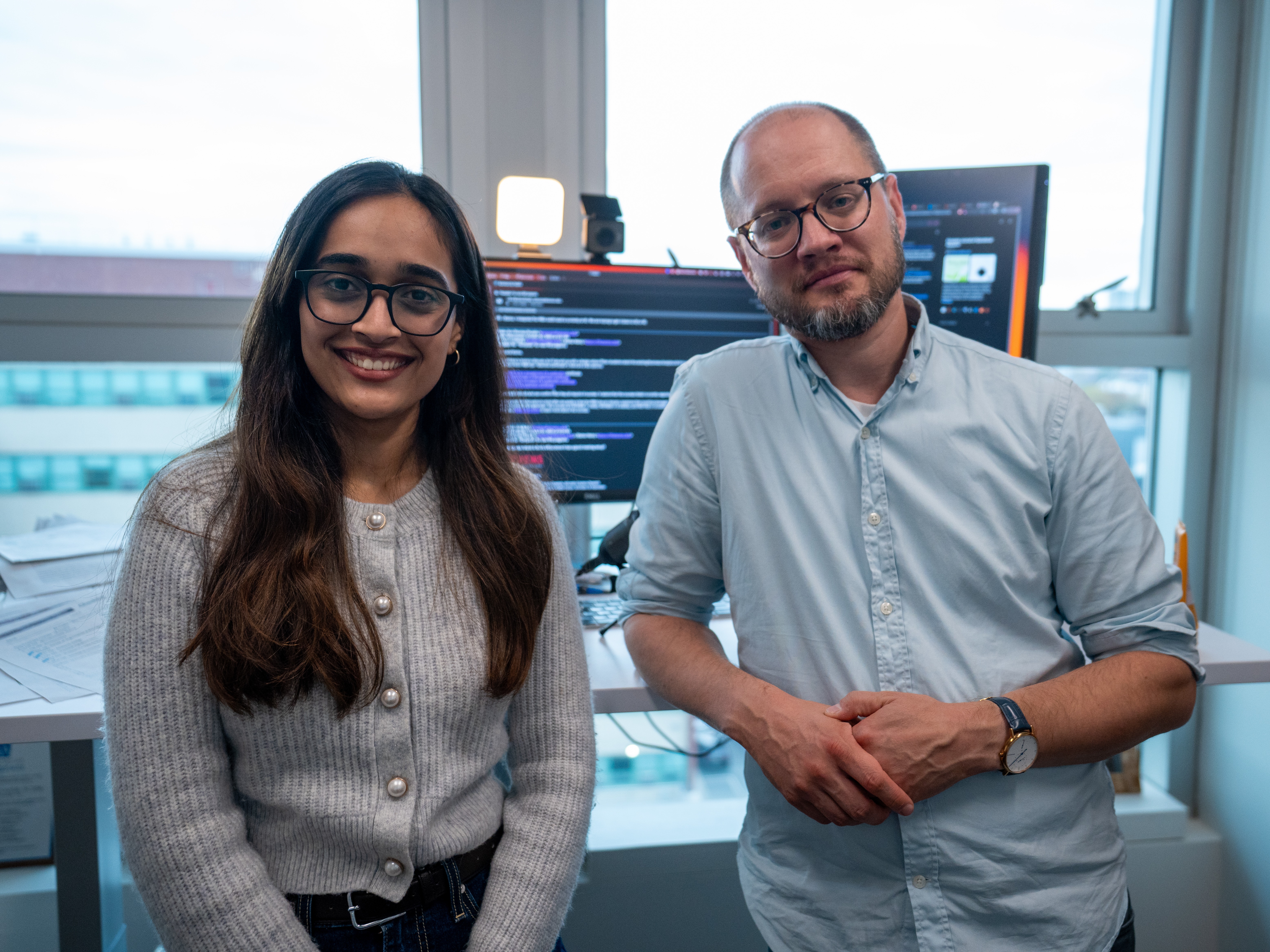 A young woman with long dark hair and glasses in a beige cardigan stands next to a bald man with glasses and beard wearing a blue button-down shirt in a modern office. Computer monitors displaying code are visible in the background along with windows.