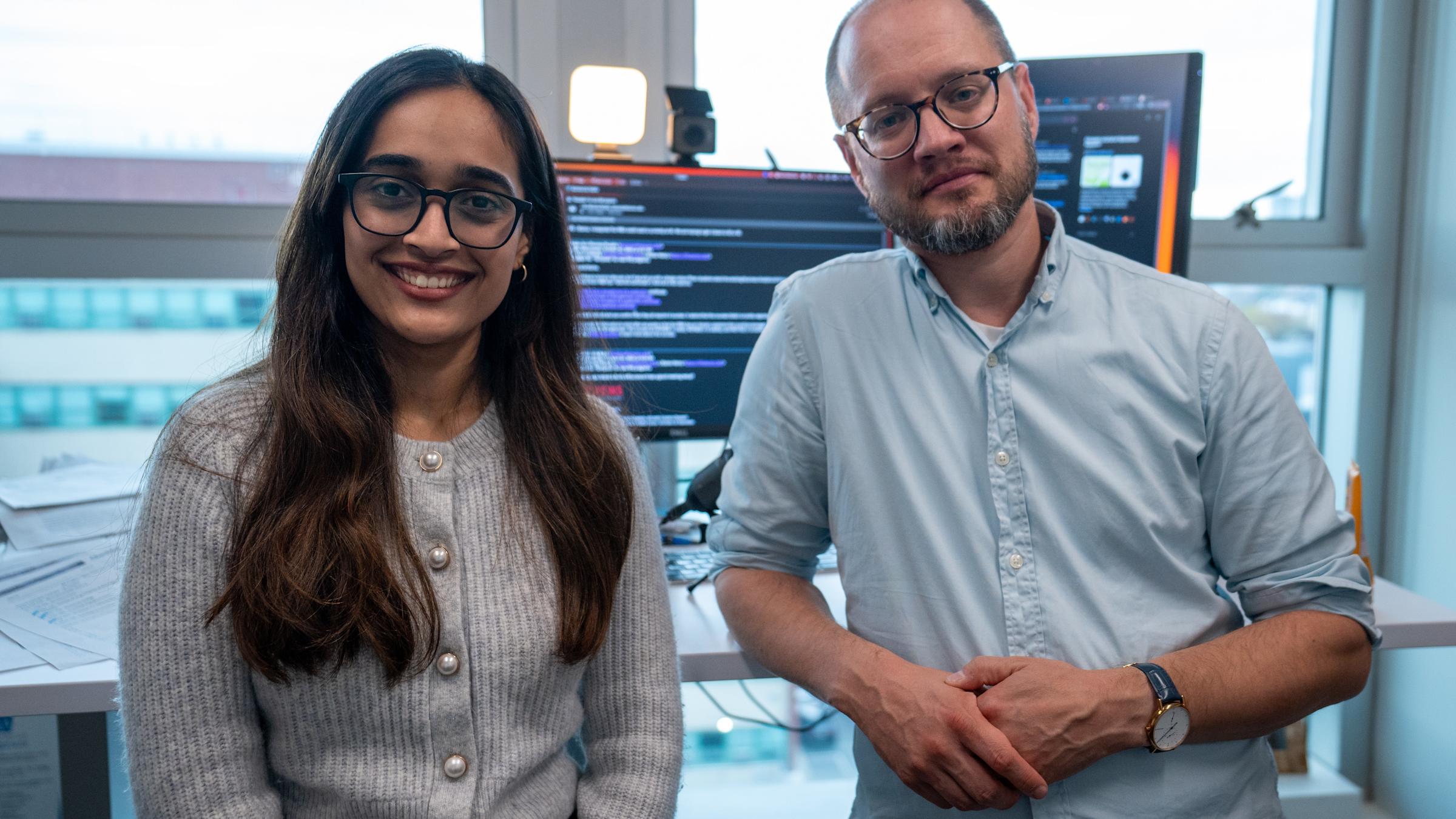 A young woman with long dark hair and glasses in a beige cardigan stands next to a bald man with glasses and beard wearing a blue button-down shirt in a modern office. Computer monitors displaying code are visible in the background along with windows.