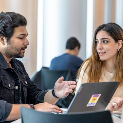 Two graduate students converse in front of their laptops.