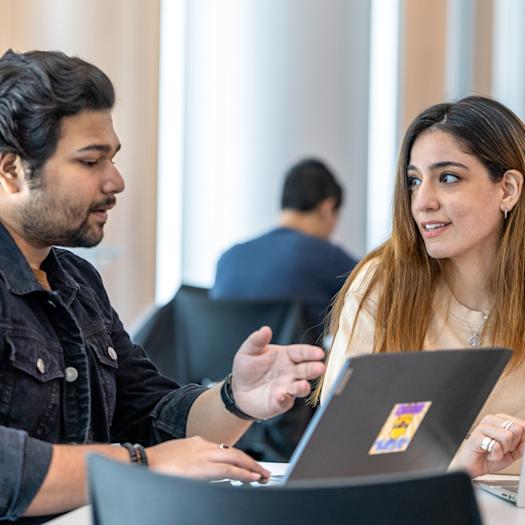 Two graduate students converse in front of their laptops.