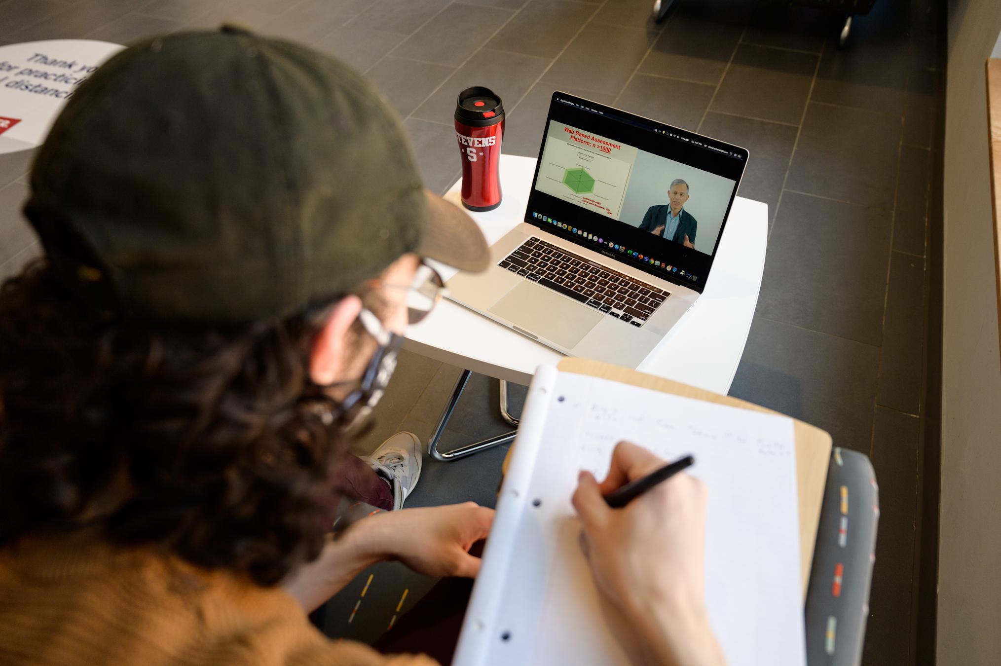 Student in ball cap writes notes while viewing Stevens lecture on laptop