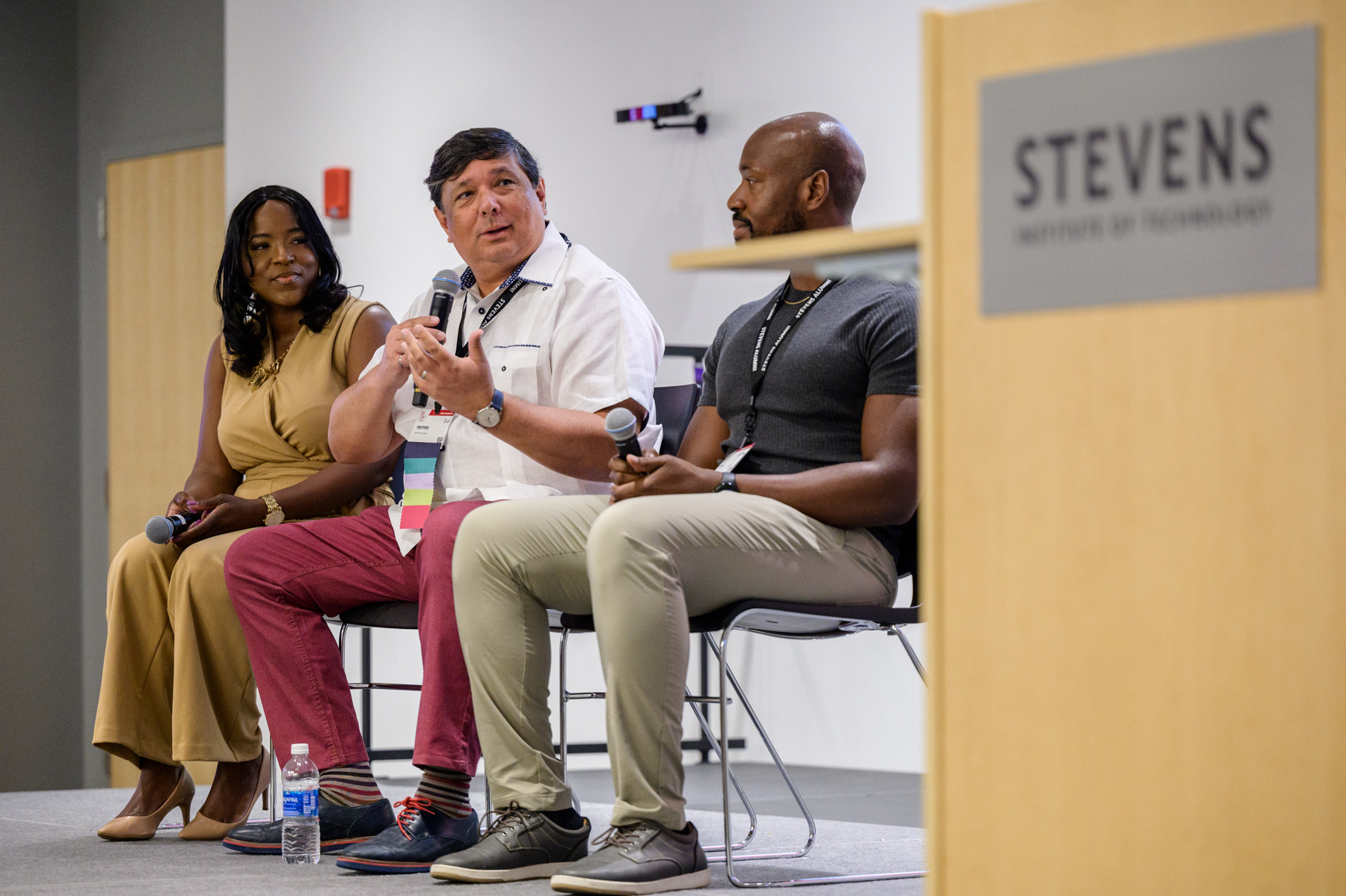 Three panelists seated onstage at a Stevens Institute of Technology event, speaking into microphones during a discussion. A podium with the Stevens logo is visible in the foreground.