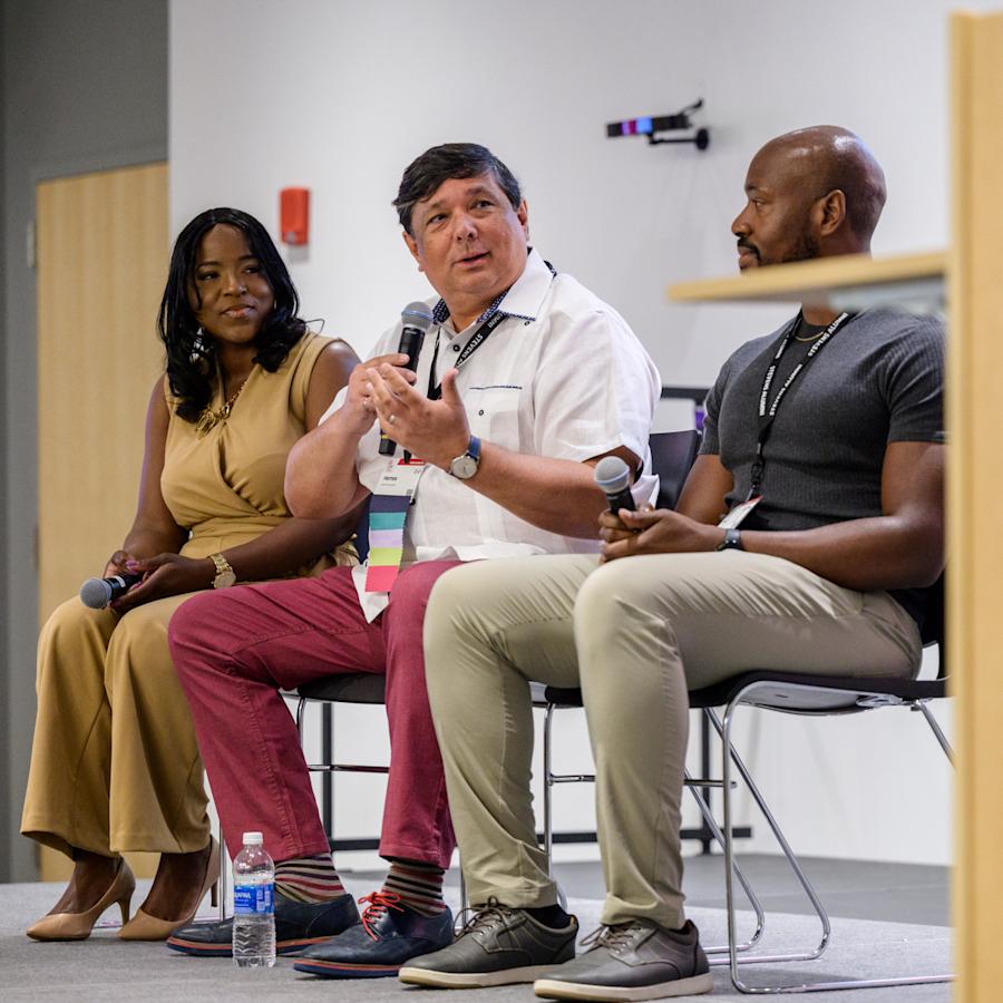 Three panelists seated onstage at a Stevens Institute of Technology event, speaking into microphones during a discussion. A podium with the Stevens logo is visible in the foreground.