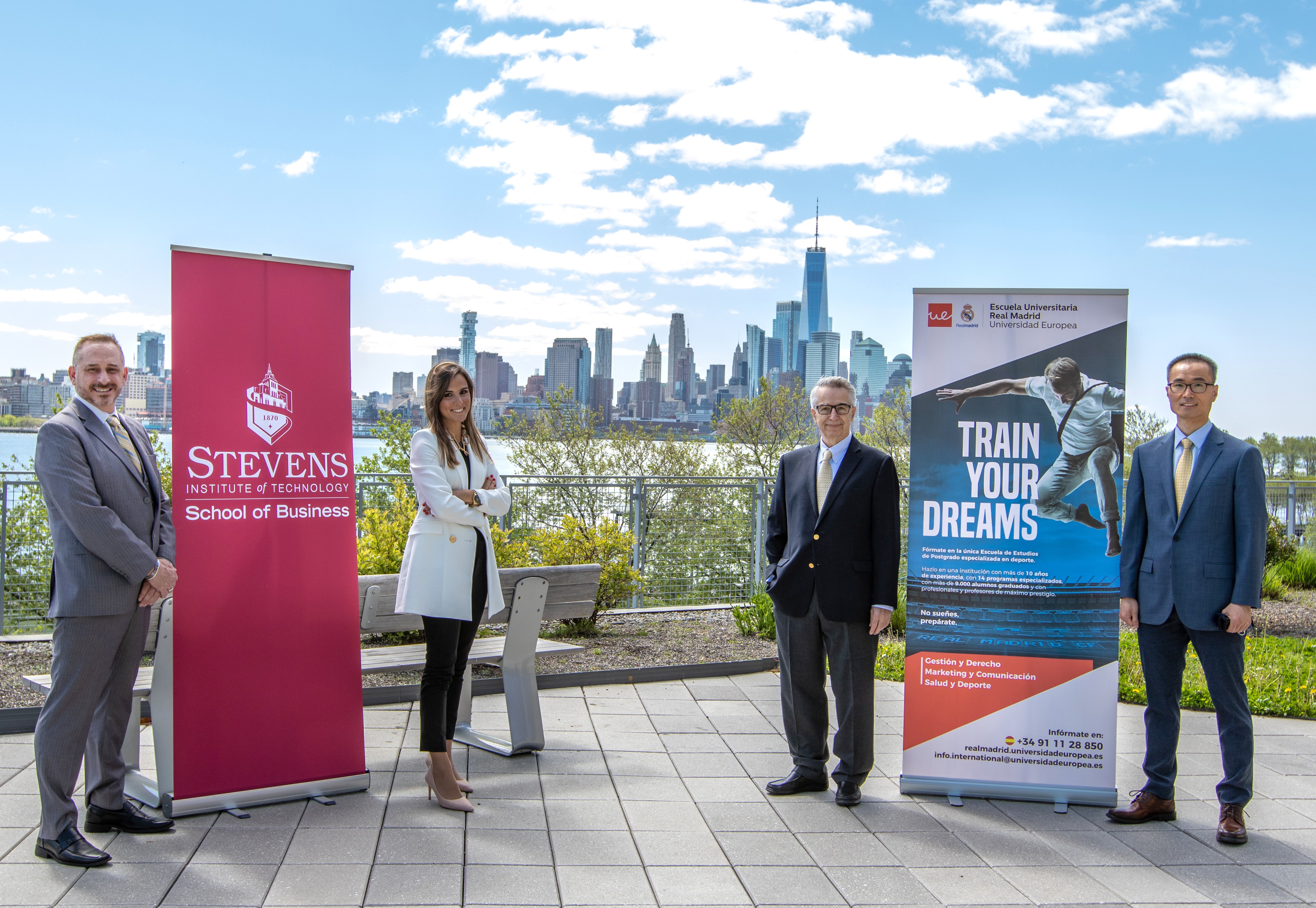 Four people standing on the Babbio patio flanking two 6-ft banners - one red with Stevens logo, representing the School of Business; the other blue multicolored representing Read Madrid. The four people from left to right are Brian Rothschild, Lura Planel
