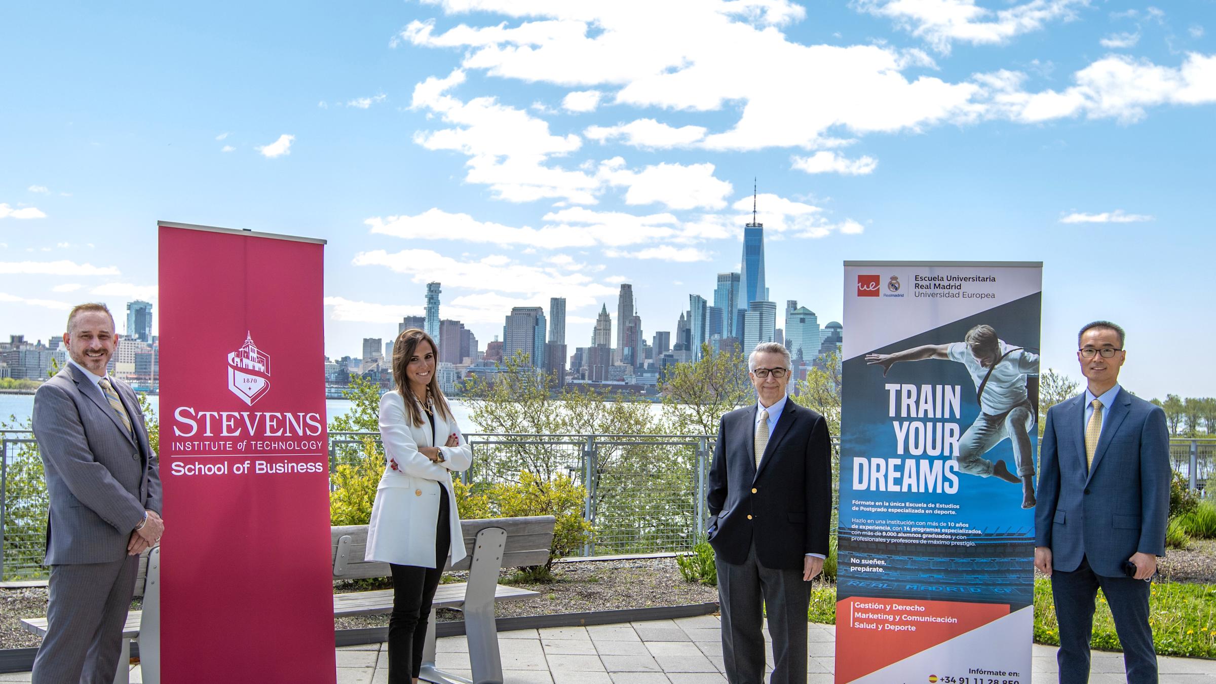 Four people standing on the Babbio patio flanking two 6-ft banners - one red with Stevens logo, representing the School of Business; the other blue multicolored representing Read Madrid. The four people from left to right are Brian Rothschild, Lura Planel
