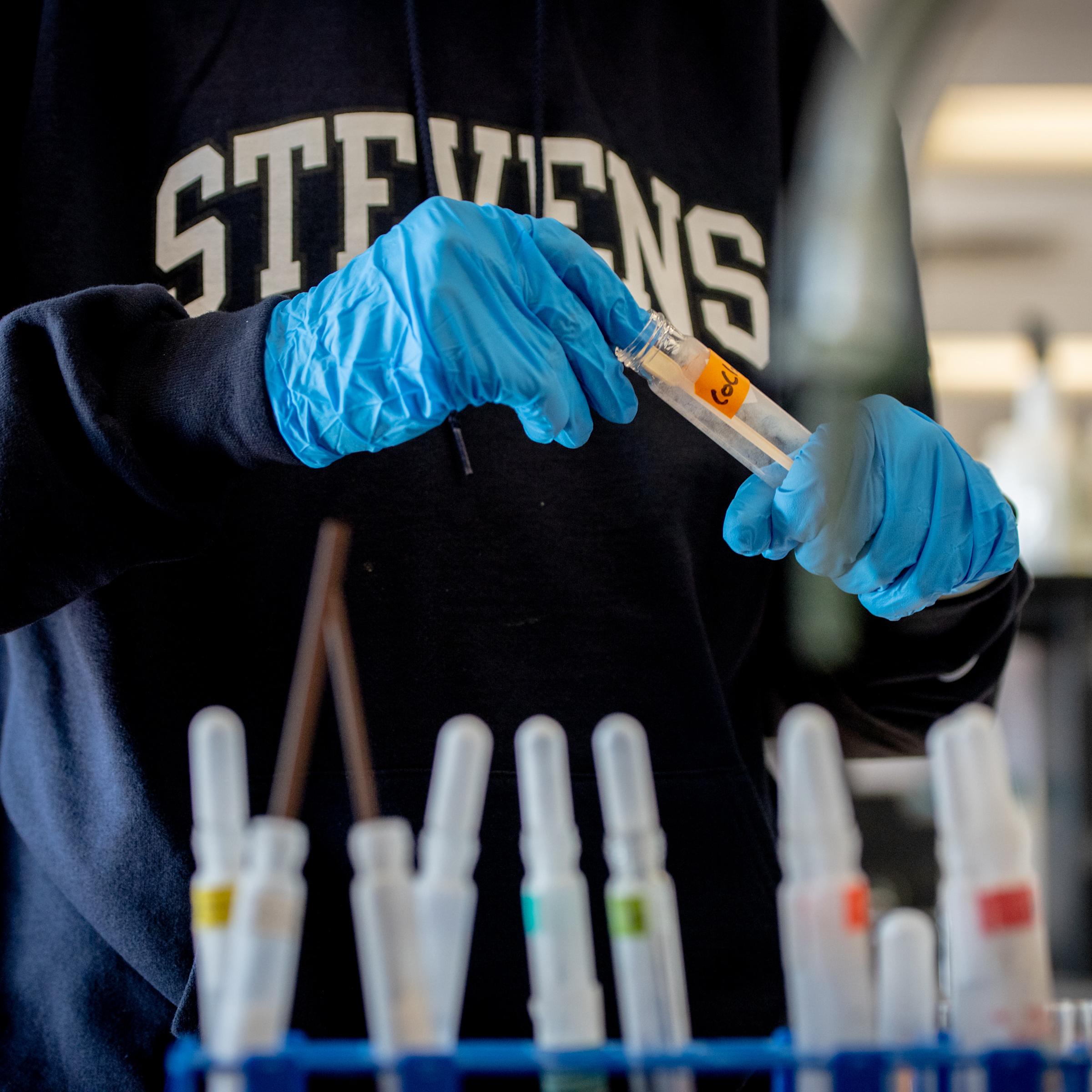 Student in a Stevens sweatshirt handling test tubes in a lab.