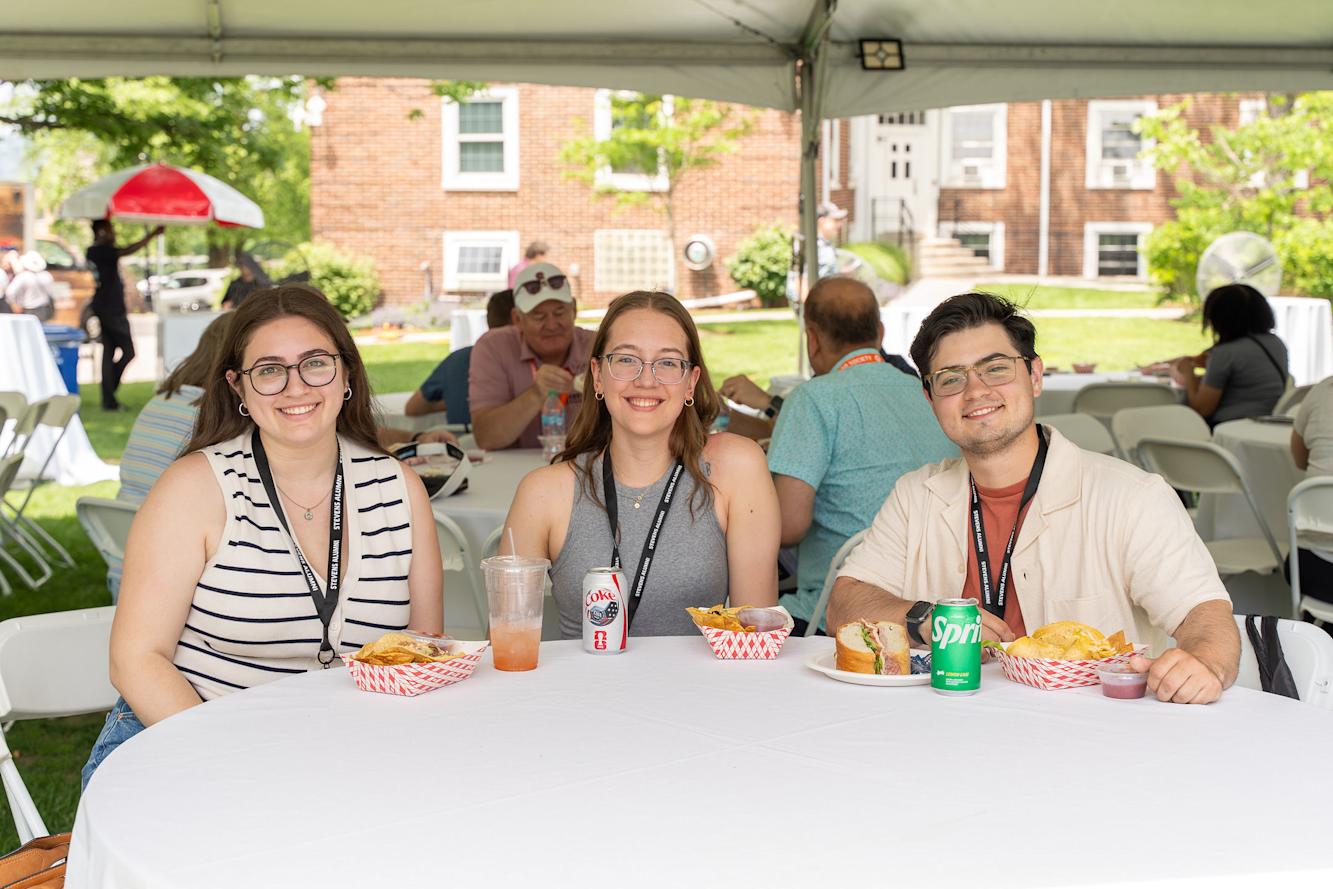 Three young alumni seated at a table outdoors during Stevens Alumni Weekend 2026