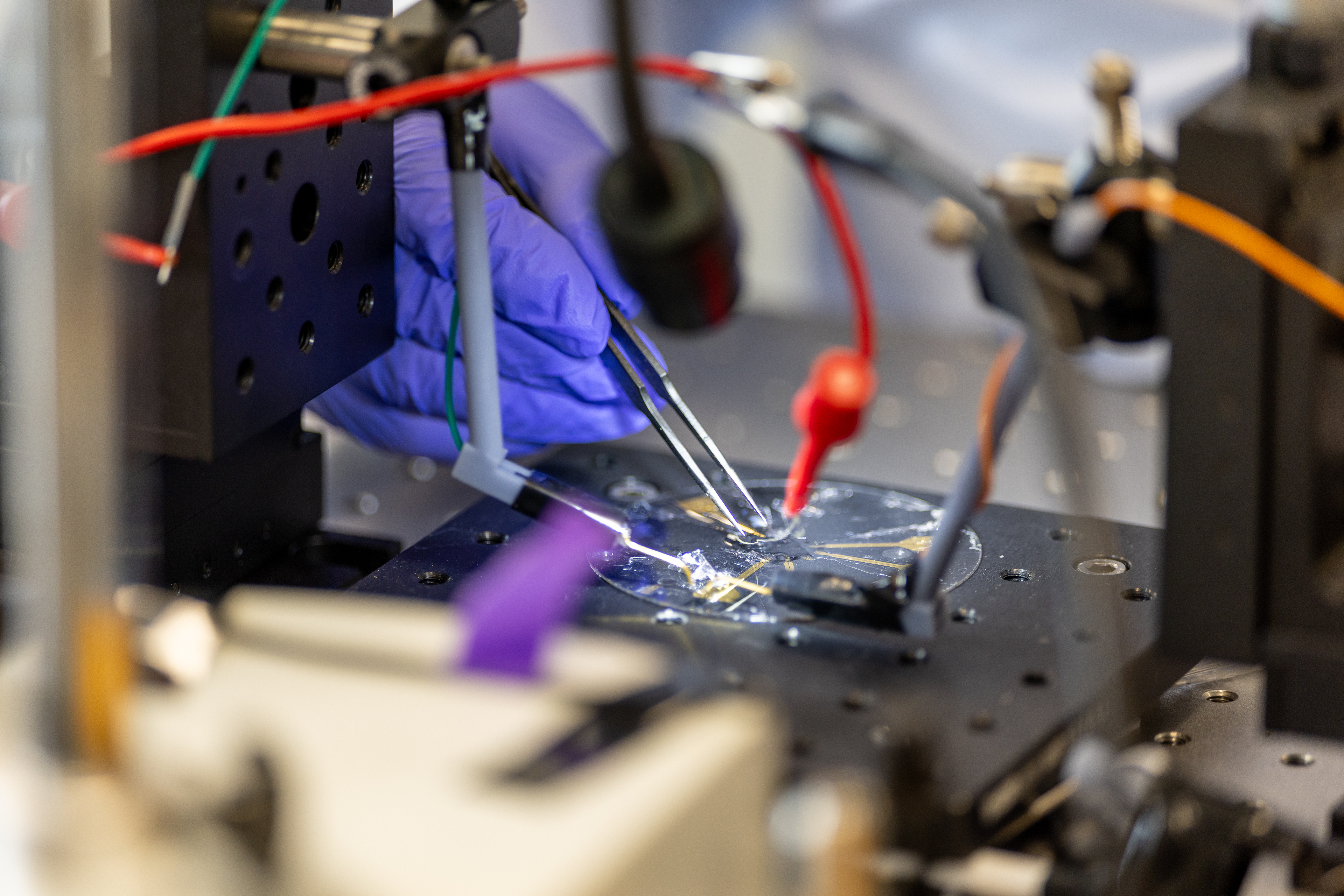 Gloved hands use tweezers to adjust components on a wired electronic setup in a lab.