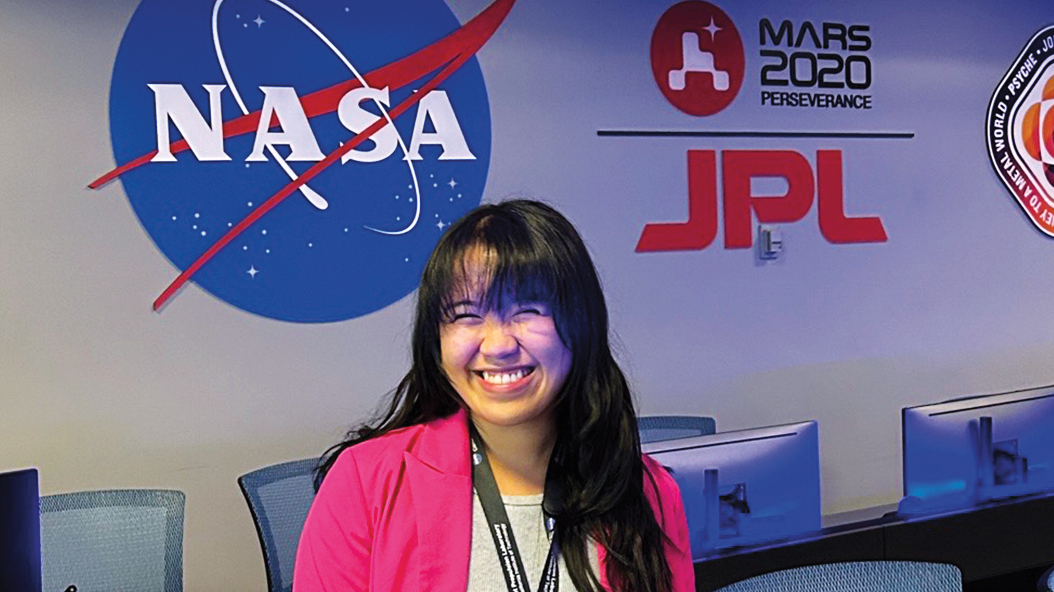 Joan Marie Tubungbunua is wearing a bright pink blazer and smiling widely. She’s in the middle of a control room where many chairs and computer monitors are lined up in two rows.