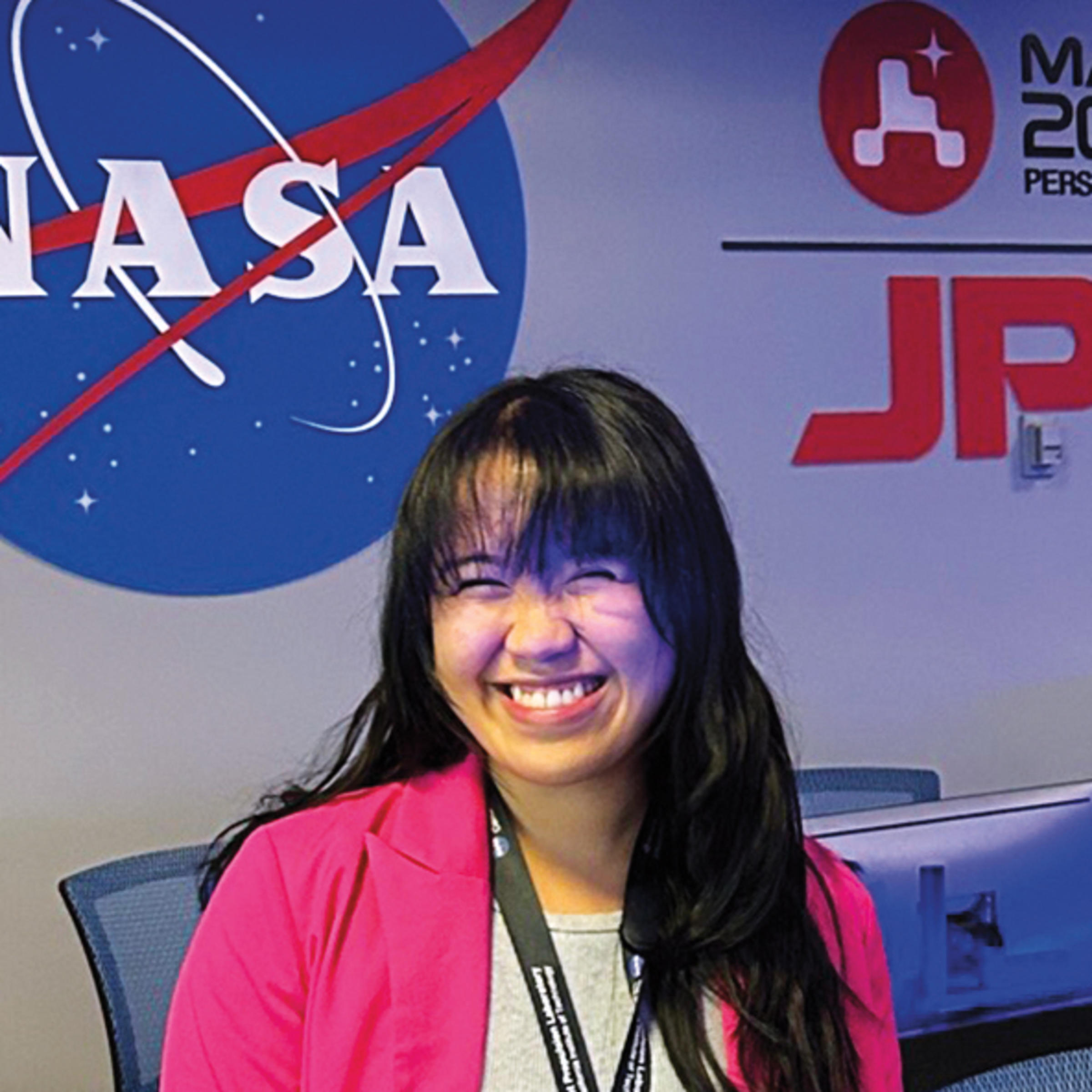 Joan Marie Tubungbunua is wearing a bright pink blazer and smiling widely. She’s in the middle of a control room where many chairs and computer monitors are lined up in two rows.