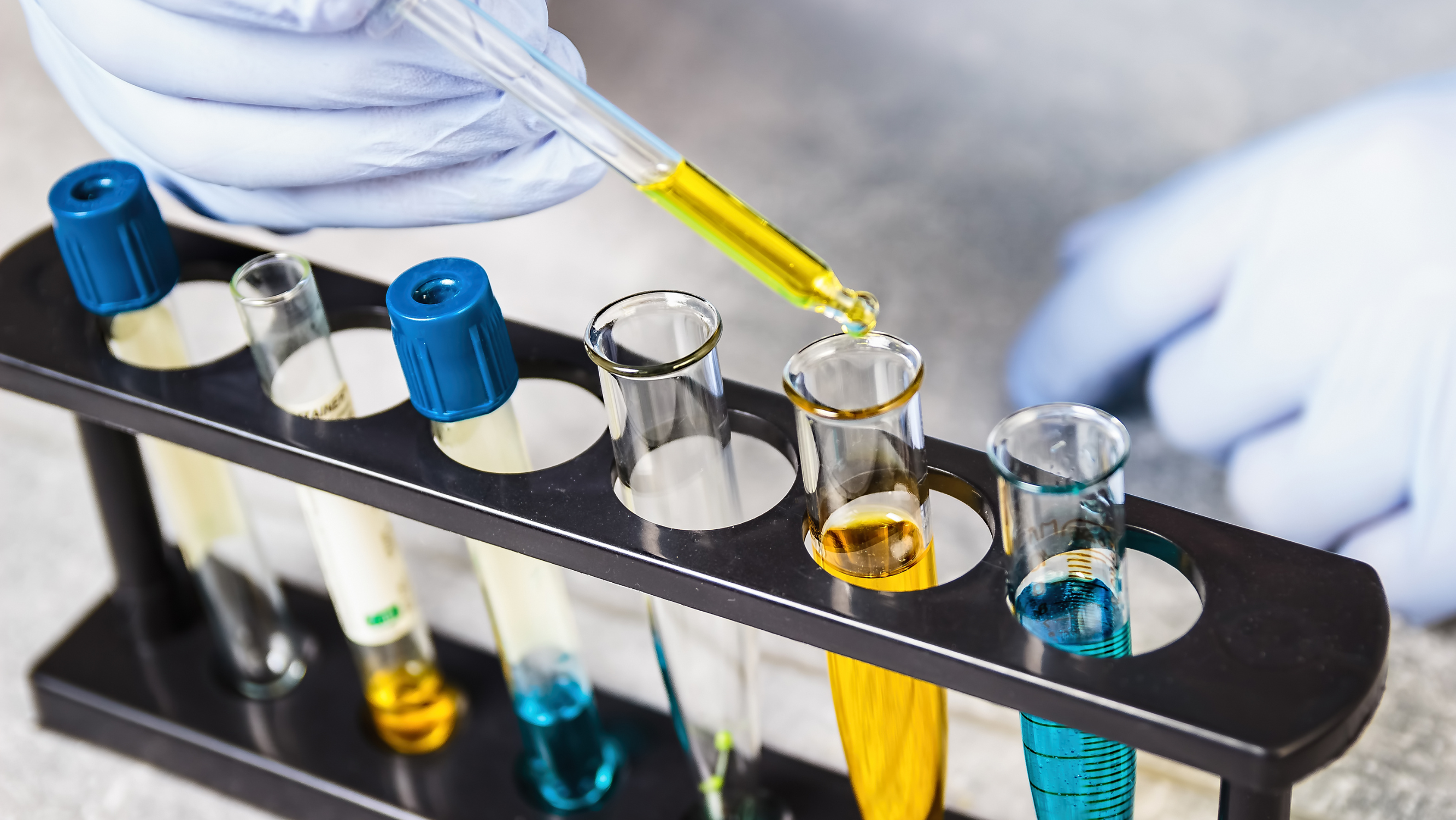 White-gloved hands pipe a liquid into a rack of test tubes containing blue and yellow liquids.