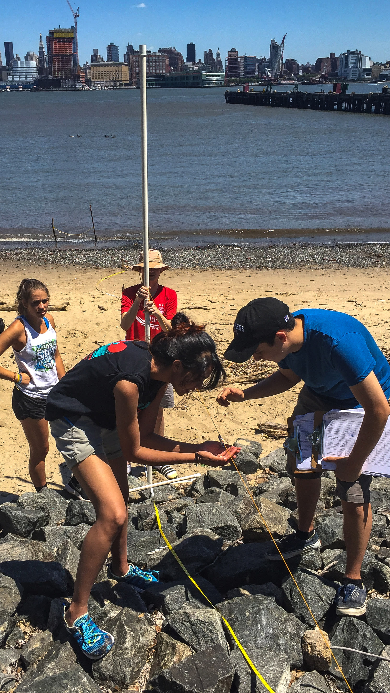 In the foreground, four Stevens students observe sand samples on the Hoboken shore with the New York City skyline in the background.