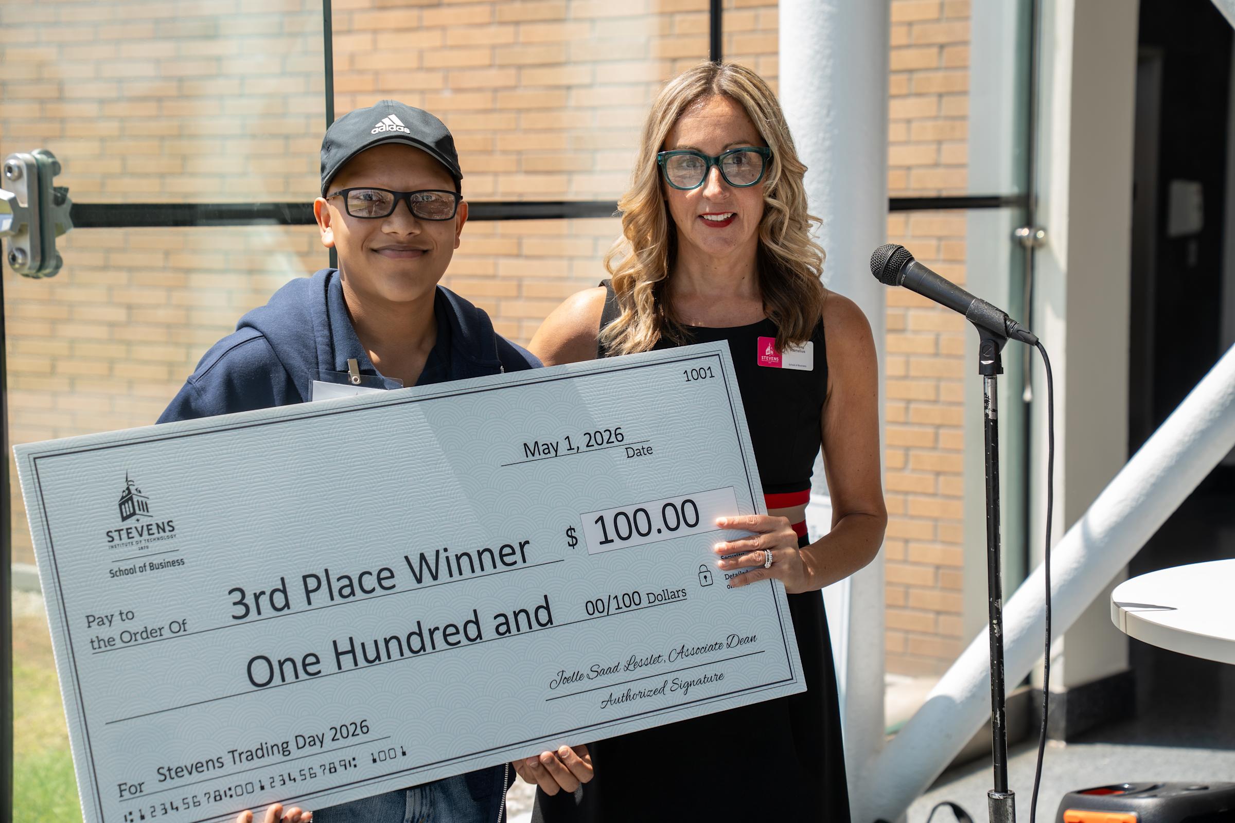A male student wearing an Adidas cap and glasses poses with a woman in black and red holding a large ceremonial check for $100 made out to "3rd Place Winner" from Stevens Institute of Technology School of Business, dated May 1, 2026.