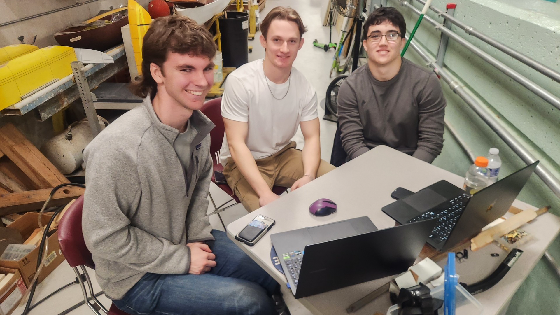 Three male Stevens undergraduate students sitting at a table with two laptops in front of them smile for the camera.