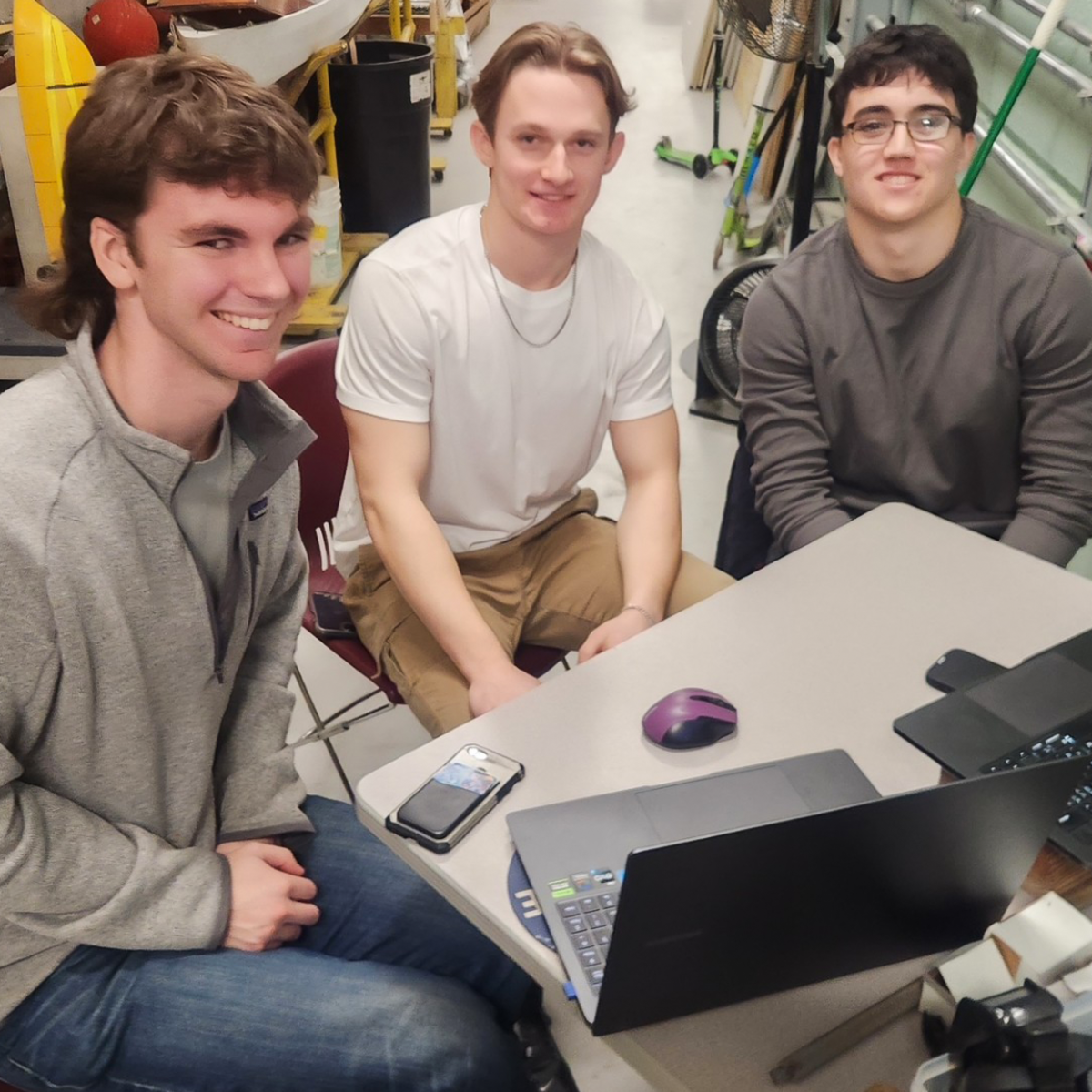 Three male Stevens undergraduate students sitting at a table with two laptops in front of them smile for the camera.