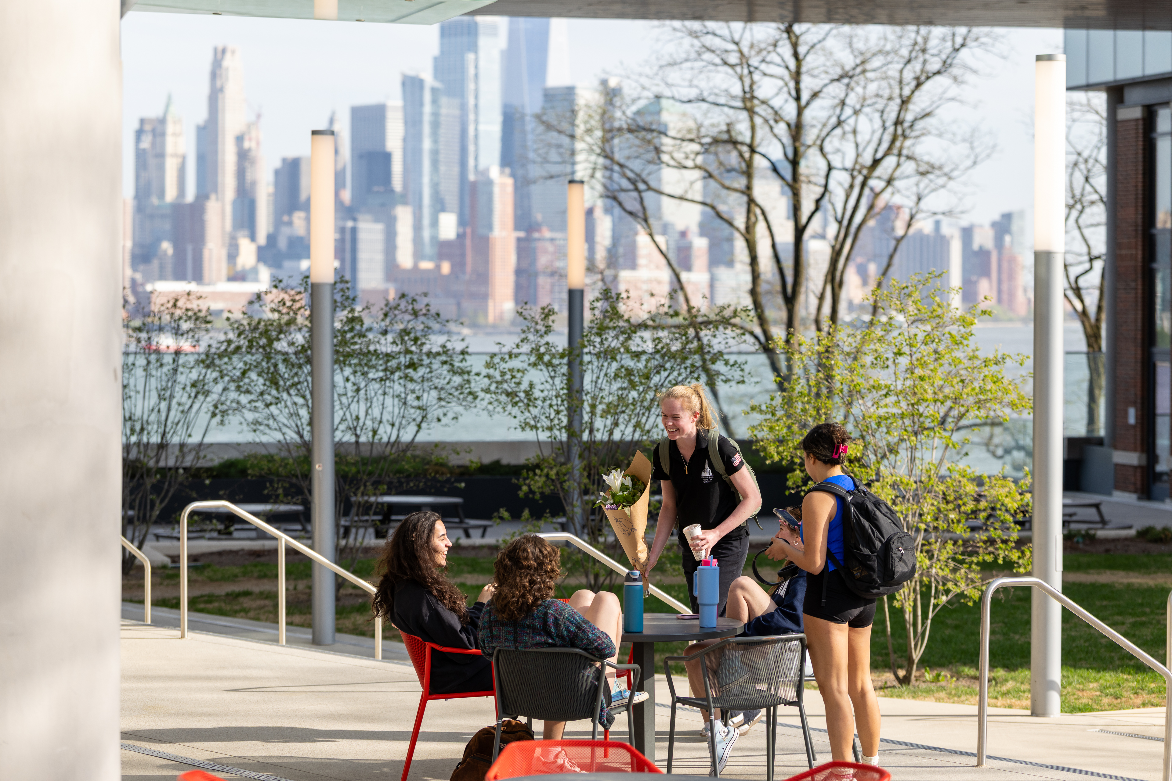 Four female students talk and laugh outside on a sunny day, with the NYC skyline in the background