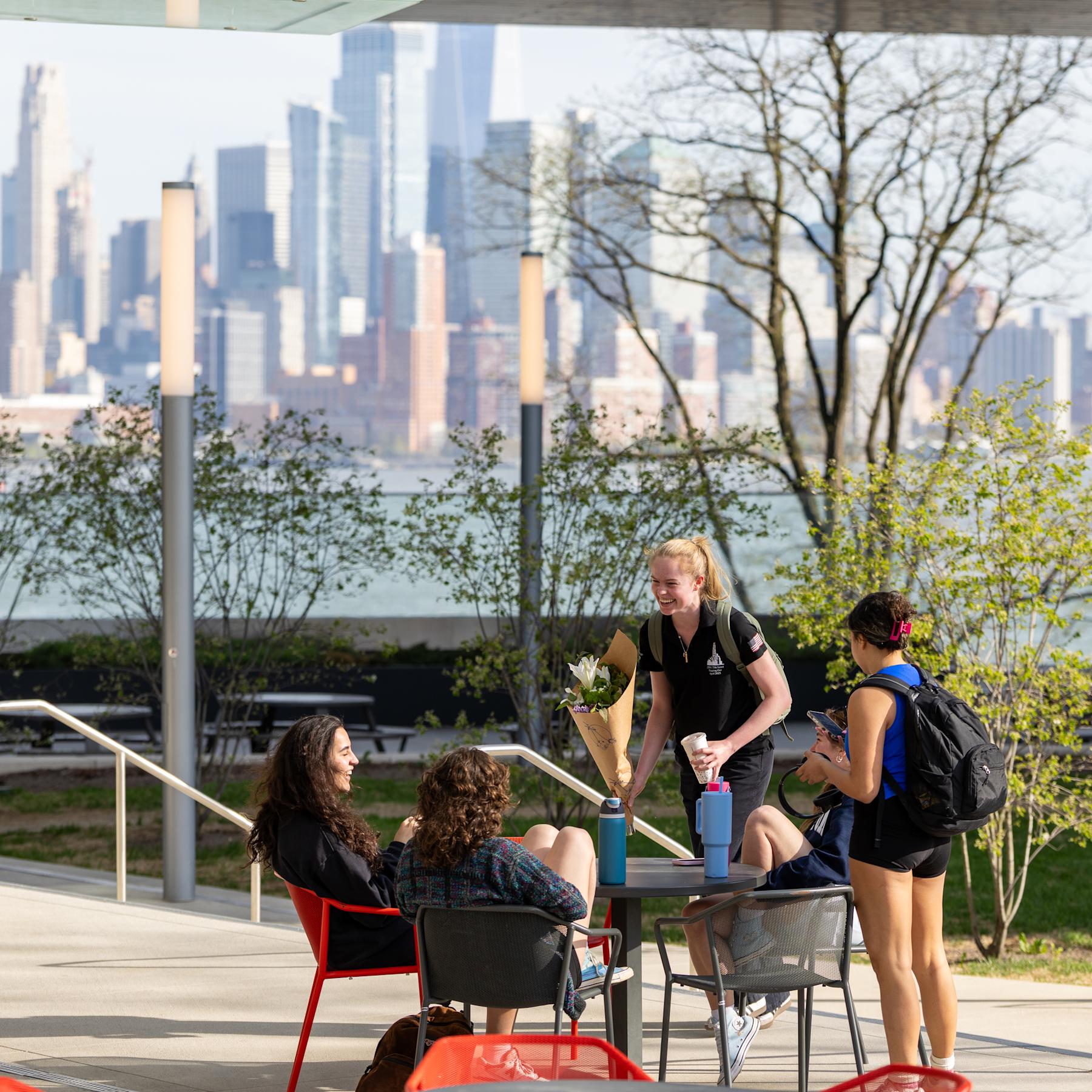 Four female students talk and laugh outside on a sunny day, with the NYC skyline in the background