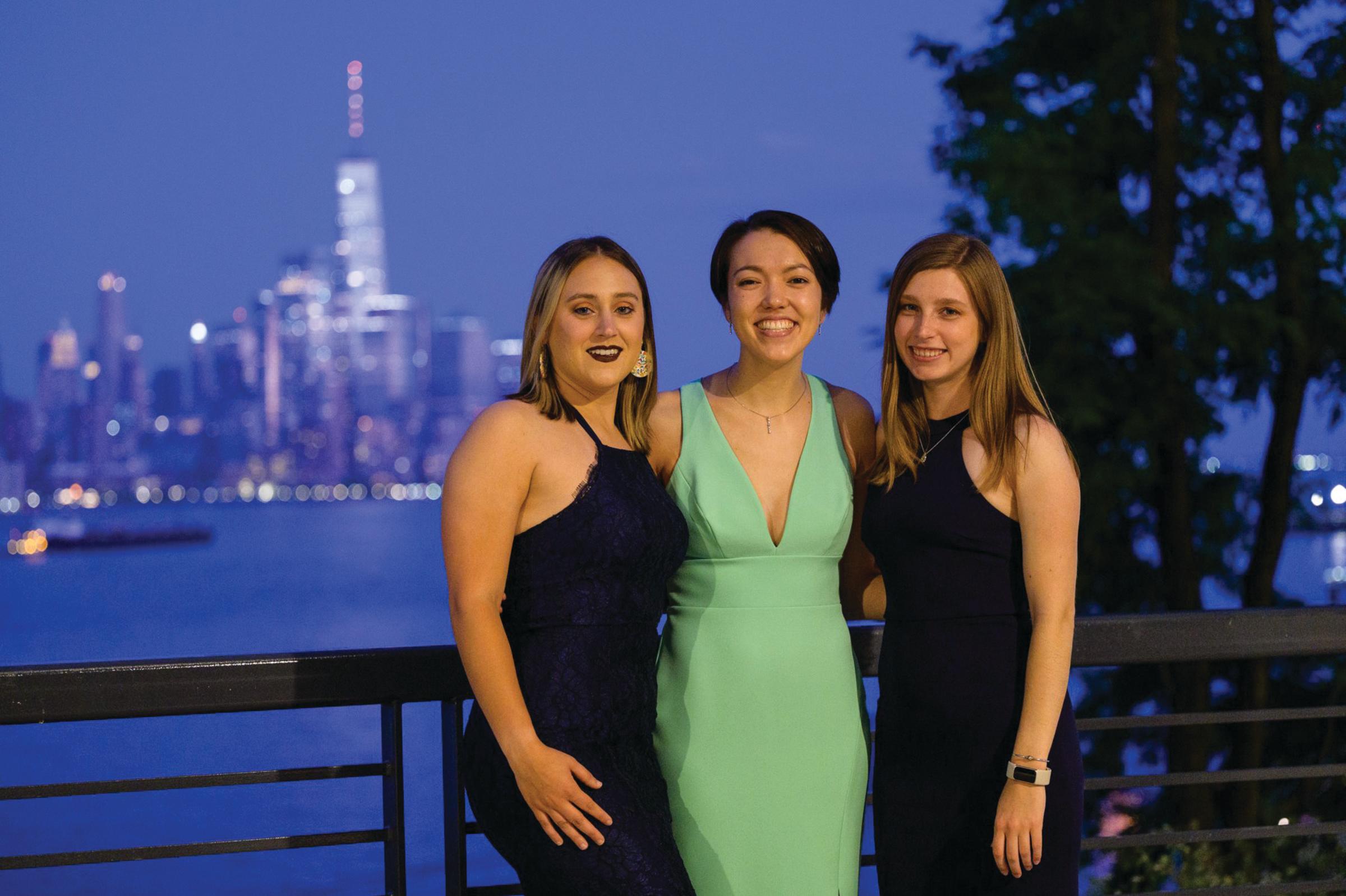 Three alumni smile for the camera in the evening with the New York skyline behind them.