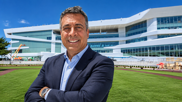 Christopher Maione ’82 stands on the grounds of the new Belmont Park racetrack in blue blazer, with the modern glass and white building rising behind him.