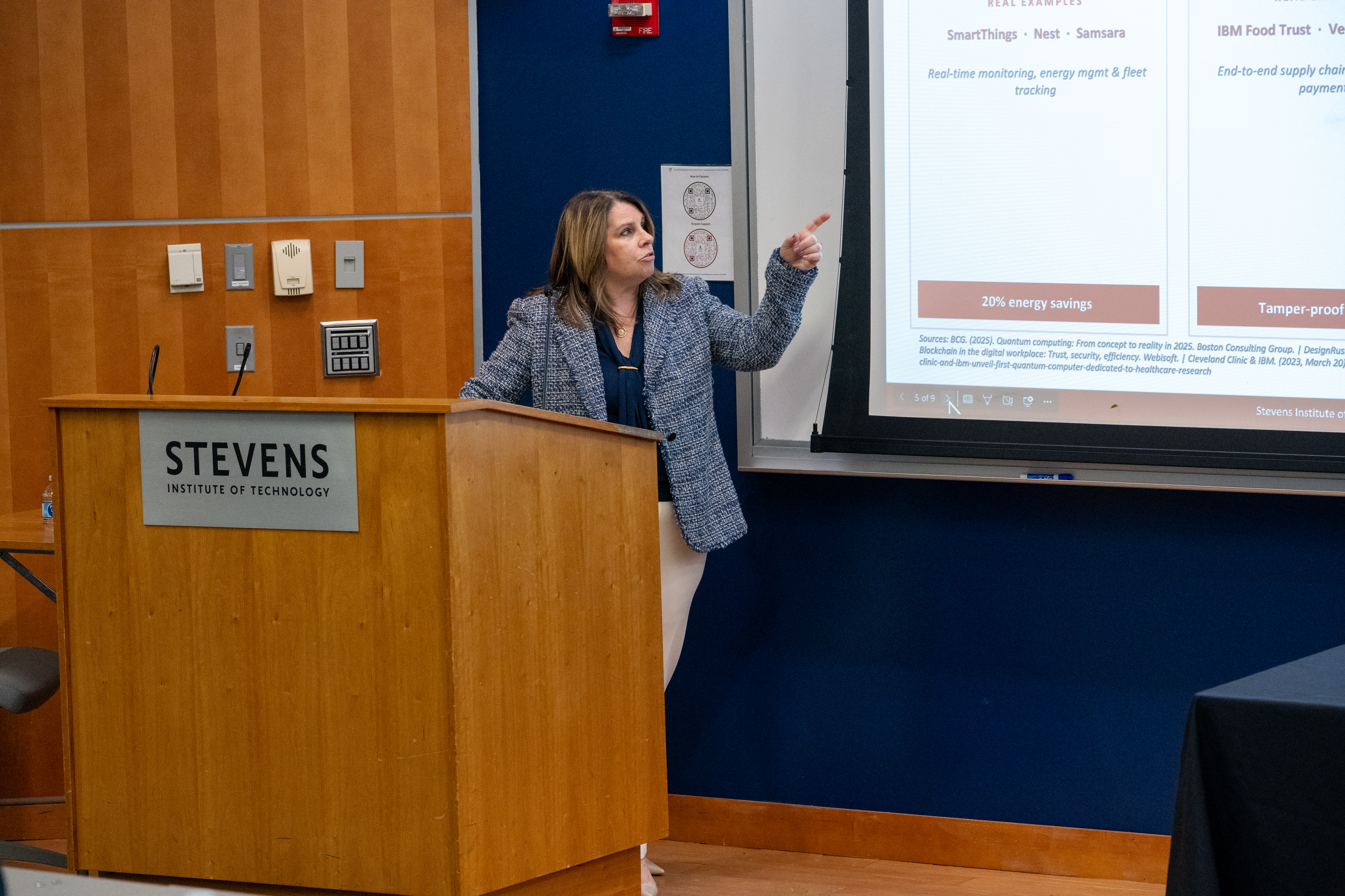 A woman in a patterned blazer presents from a wooden podium bearing the Stevens Institute of Technology logo, gesturing toward a projected slide about energy savings and blockchain applications including SmartThings, Nest, and IBM Food Trust examples.