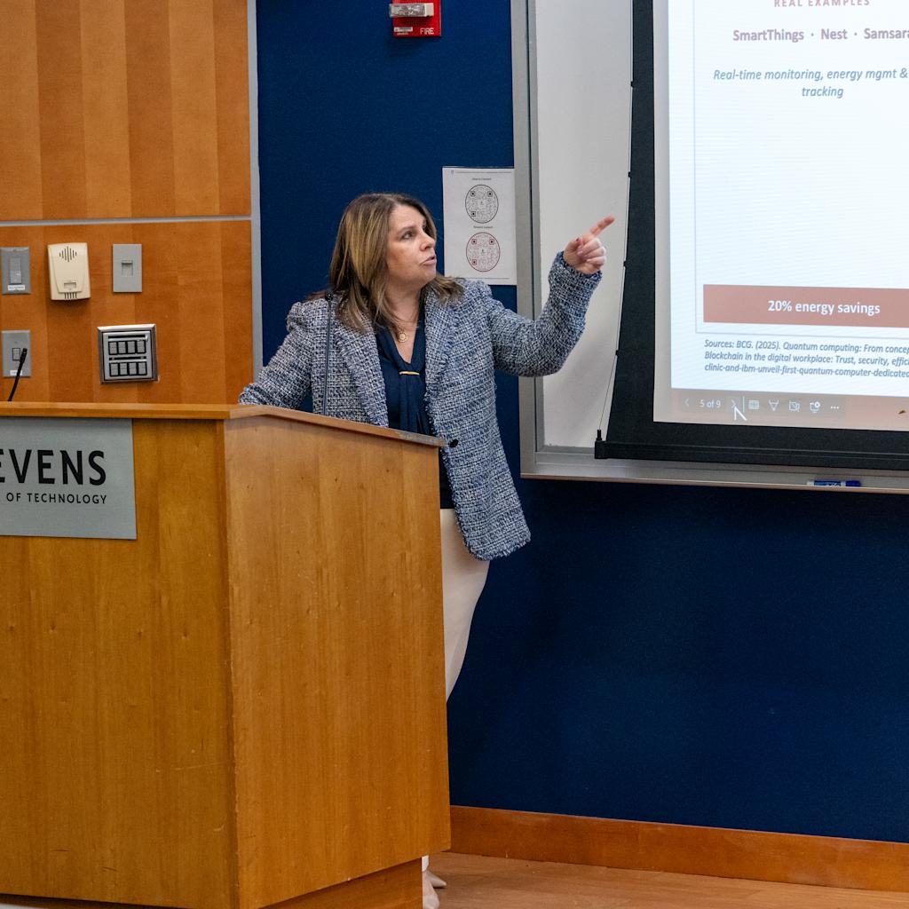 A woman in a patterned blazer presents from a wooden podium bearing the Stevens Institute of Technology logo, gesturing toward a projected slide about energy savings and blockchain applications including SmartThings, Nest, and IBM Food Trust examples.