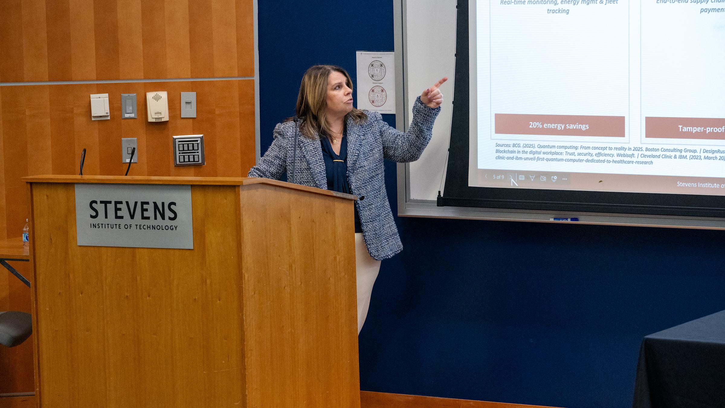 A woman in a patterned blazer presents from a wooden podium bearing the Stevens Institute of Technology logo, gesturing toward a projected slide about energy savings and blockchain applications including SmartThings, Nest, and IBM Food Trust examples.