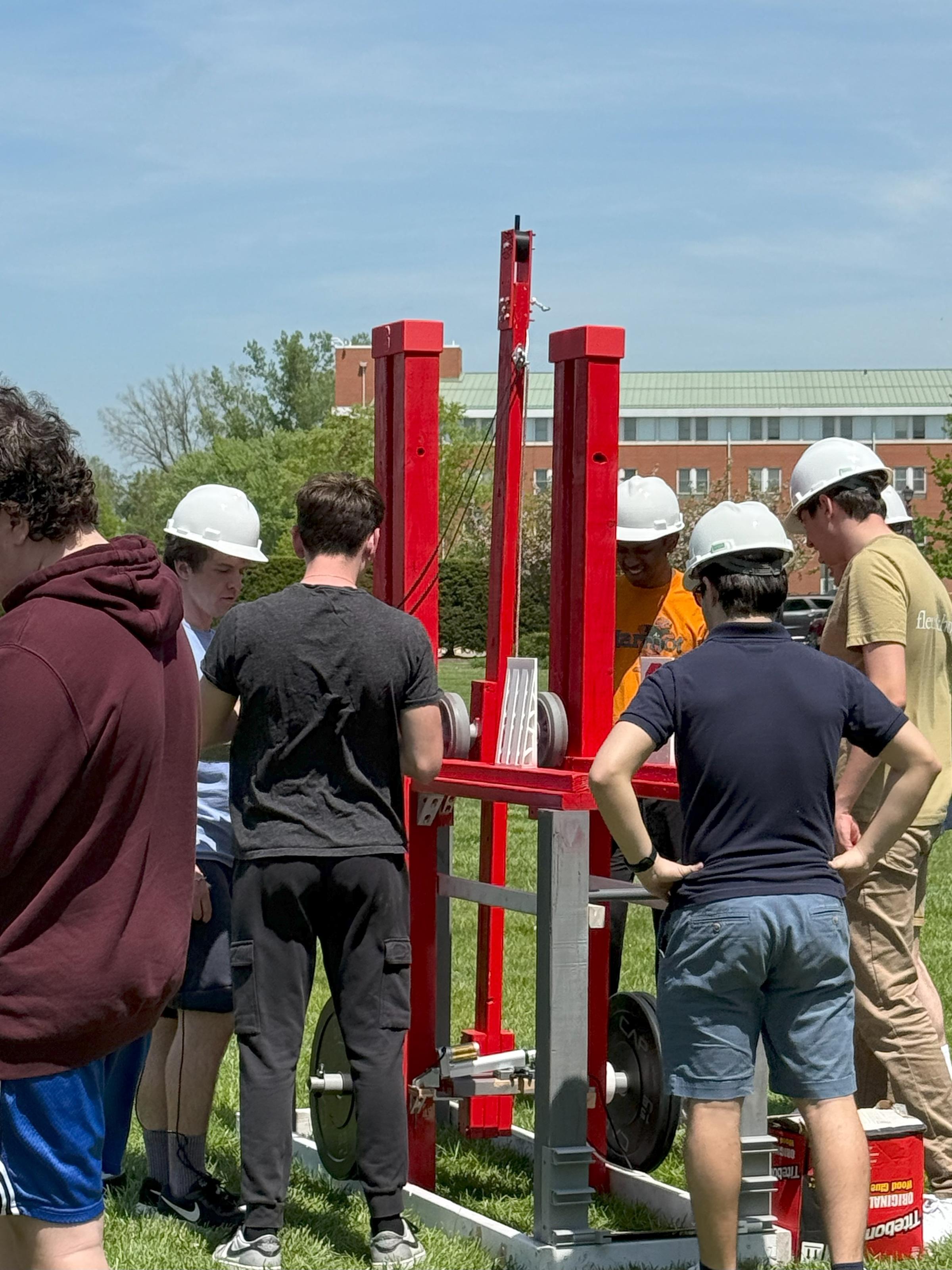 A group of students wearing hard hats stand around their trebuchet machine.